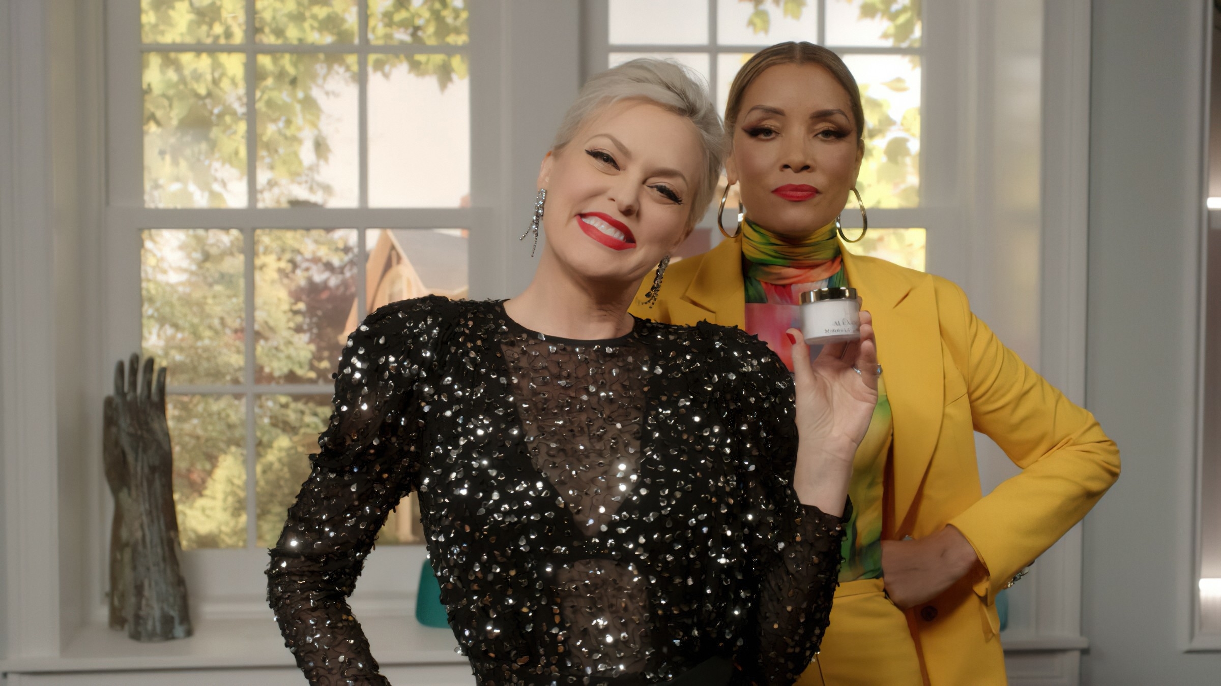 Alexis in a sparkly dress holding a makeup product, in front of woman in colorful suit