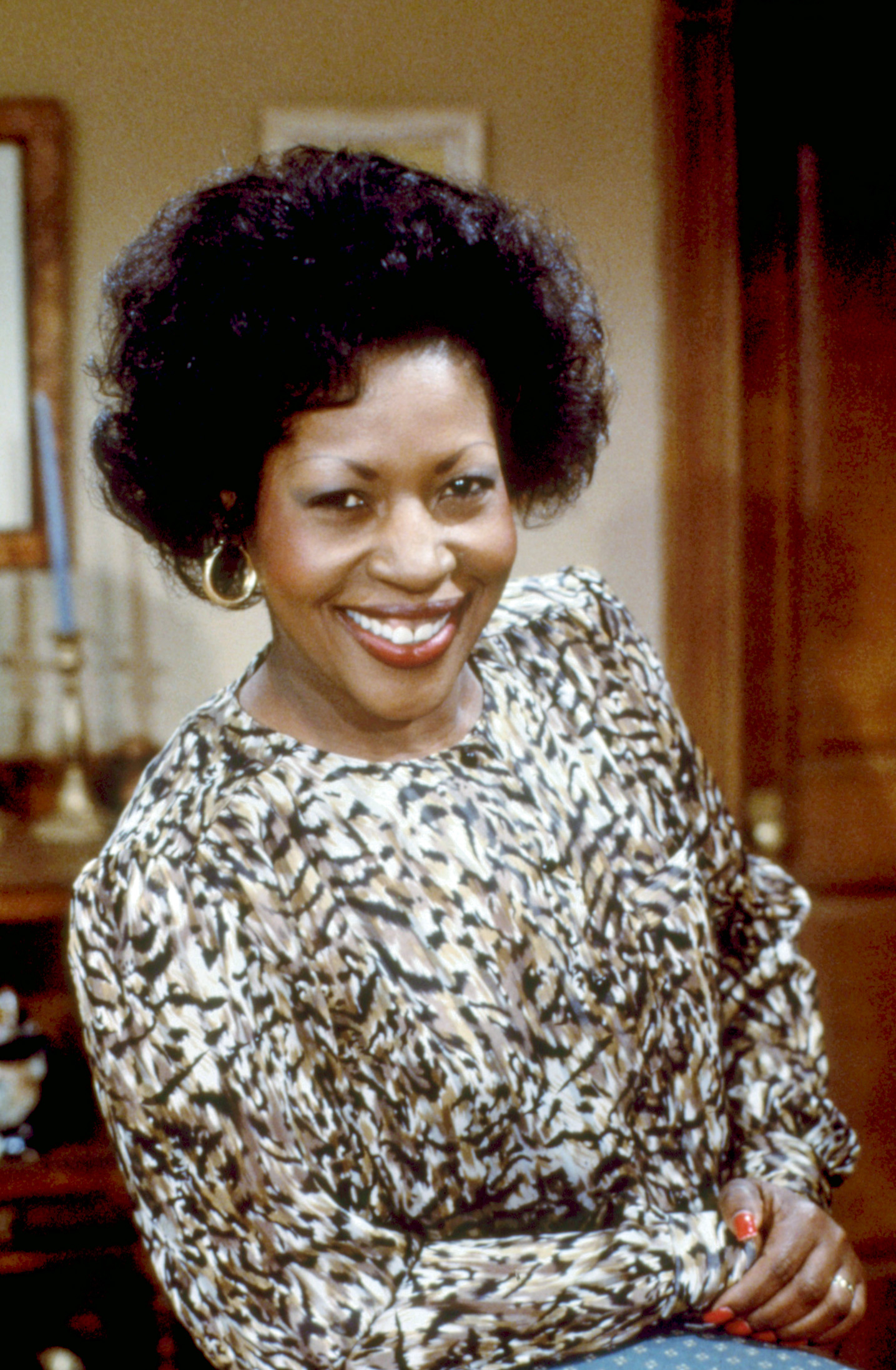 Harriette smiling while wearing a patterned blouse with puffed sleeves and hoop earrings