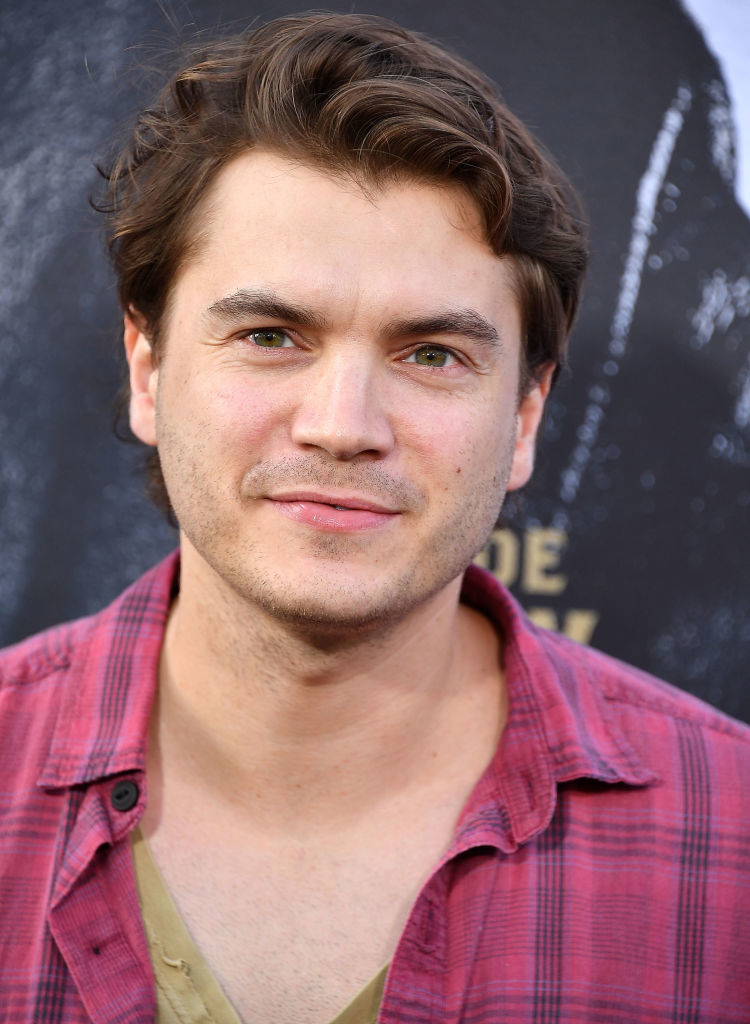 Emile Hirsch smiles at the camera, wearing a casual open plaid shirt over a beige t-shirt