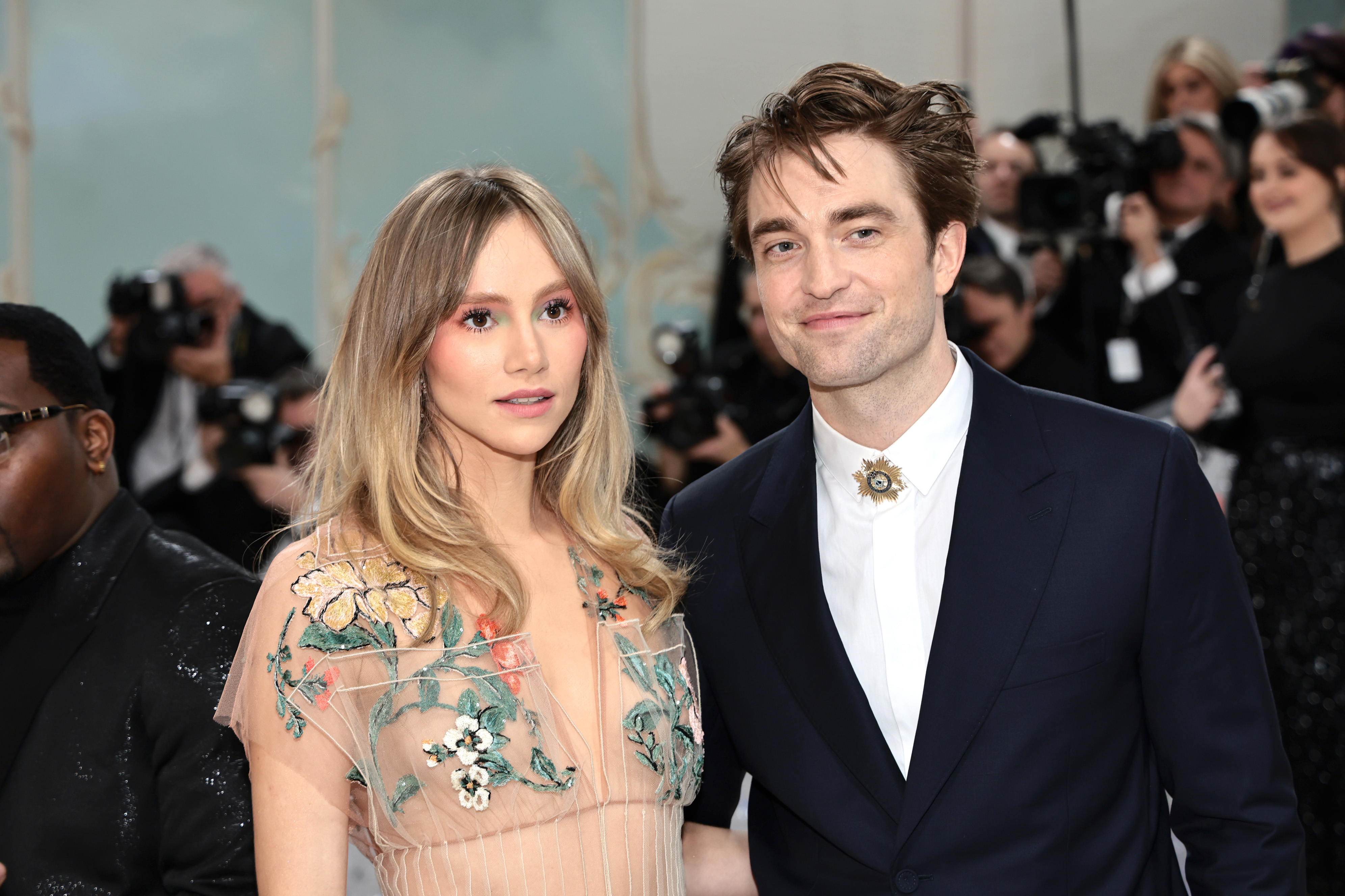 Suki Waterhouse in a floral embroidered dress and Robert Pattinson in a suit pose together at a celebrity event with photographers in the background