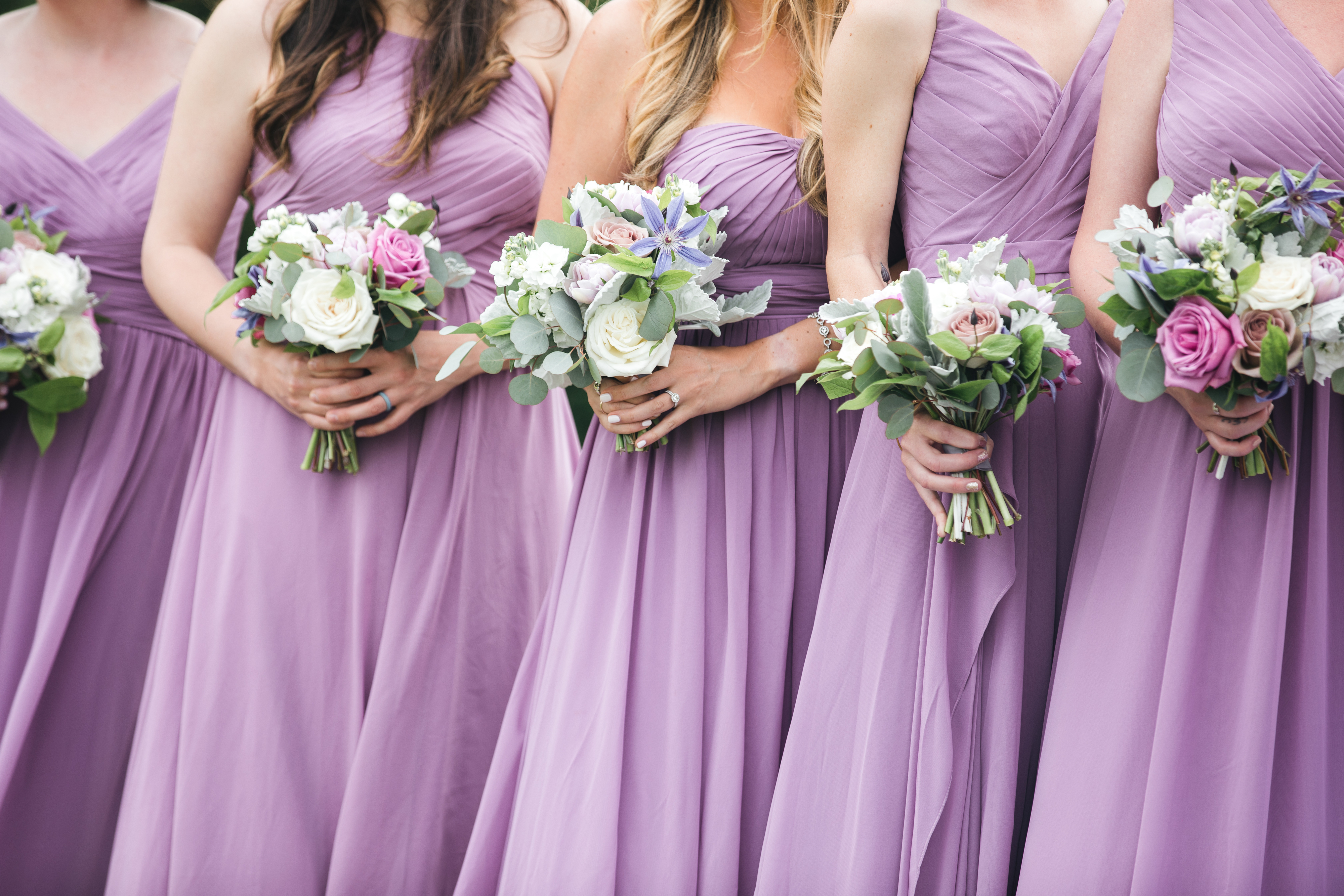 A group of bridesmaids hold bouquets while wearing floor-length, sleeveless bridesmaid dresses. Faces are not shown