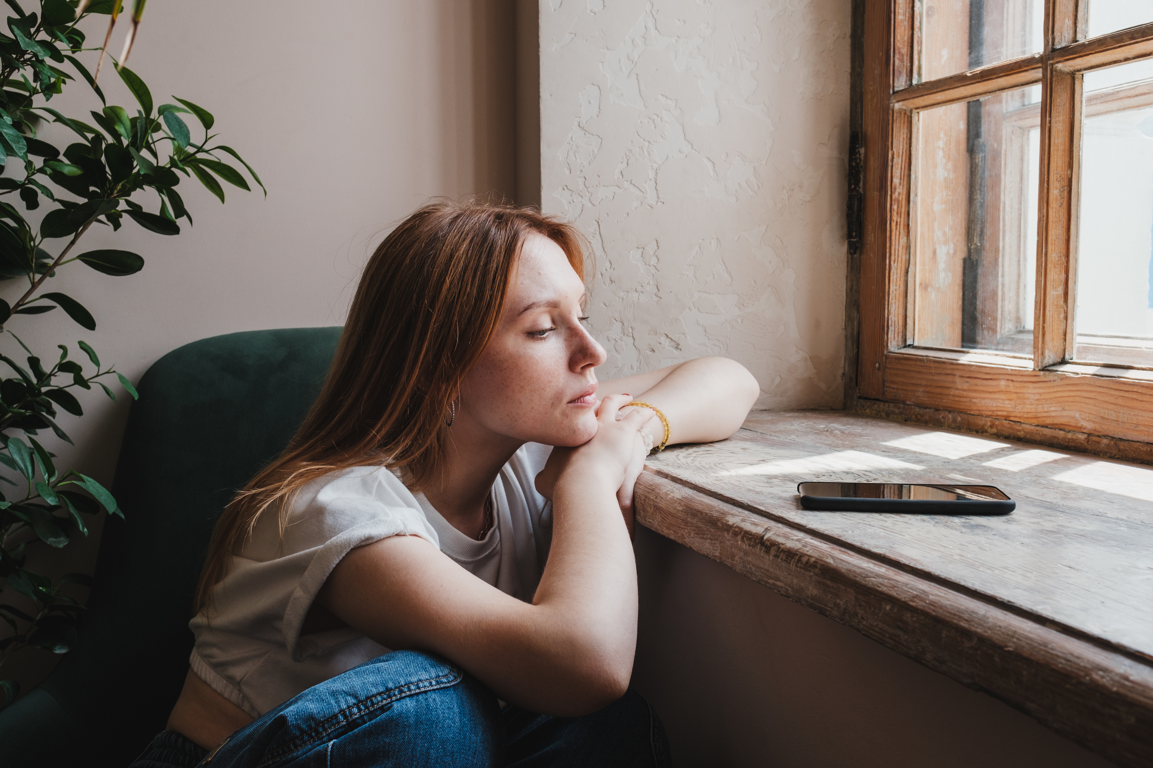 A woman with long hair sits by a window, resting her head on her arms, looking at a smartphone placed on the windowsill