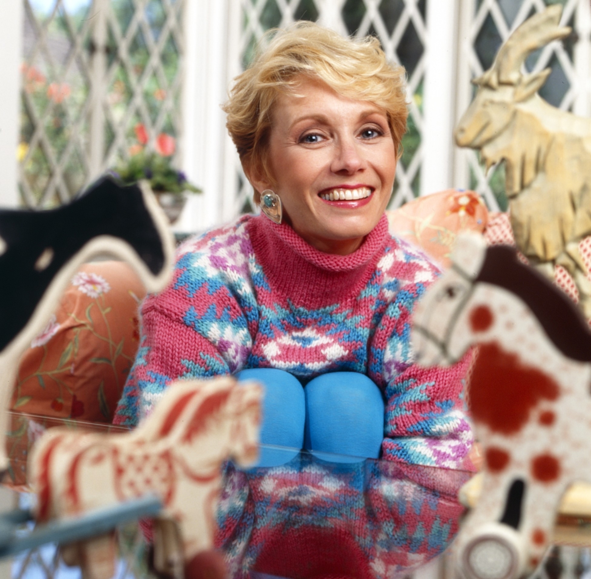 Smiling Sandy with short blonde hair, wearing a patterned sweater. She is surrounded by various wooden animal figurines, including horses and a goat