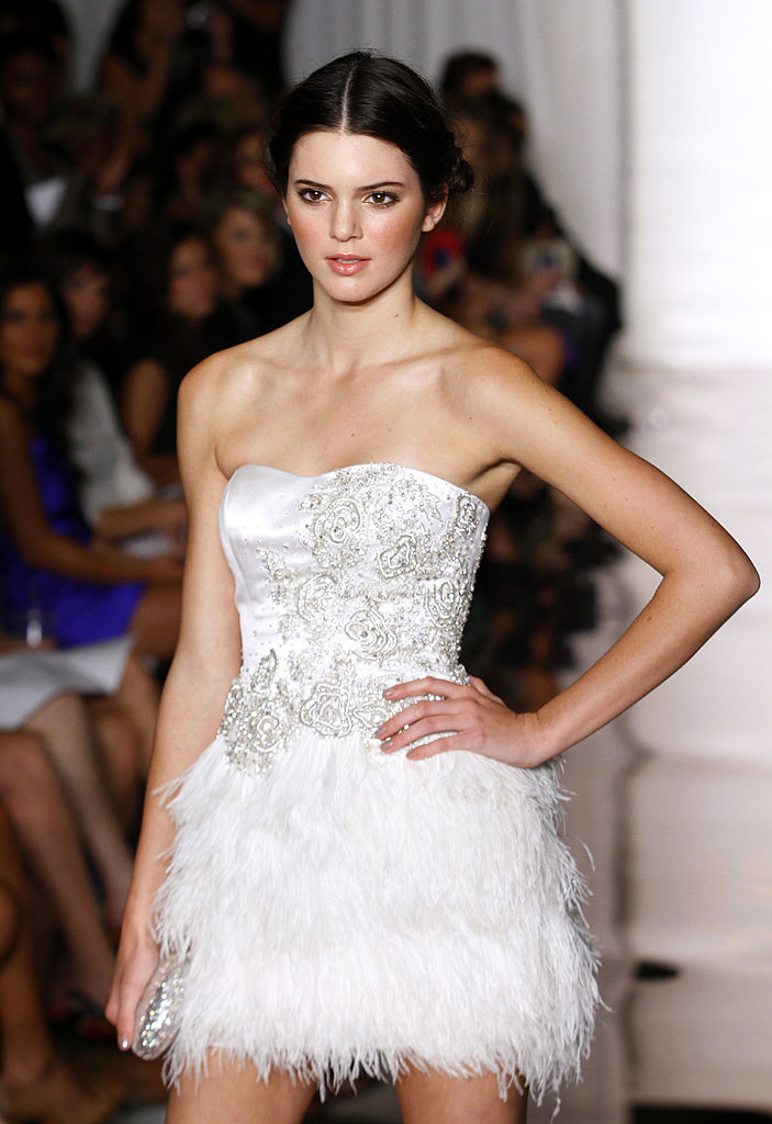 A model walks the runway in an off-the-shoulder, white, embellished mini dress with feathered details at the skirt during a fashion show