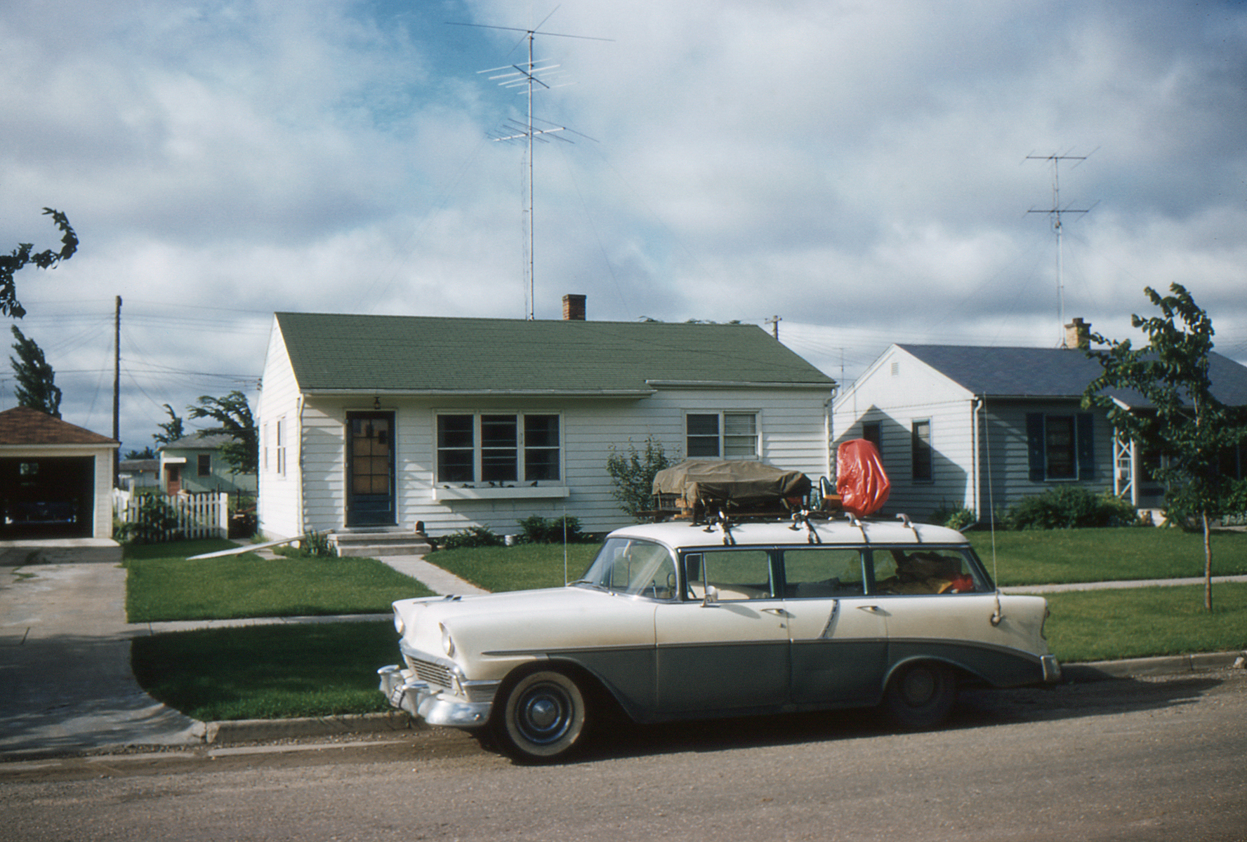 A 1956 Chevrolet station wagon with luggage on top is parked in front of a small single-story house in a suburban neighborhood
