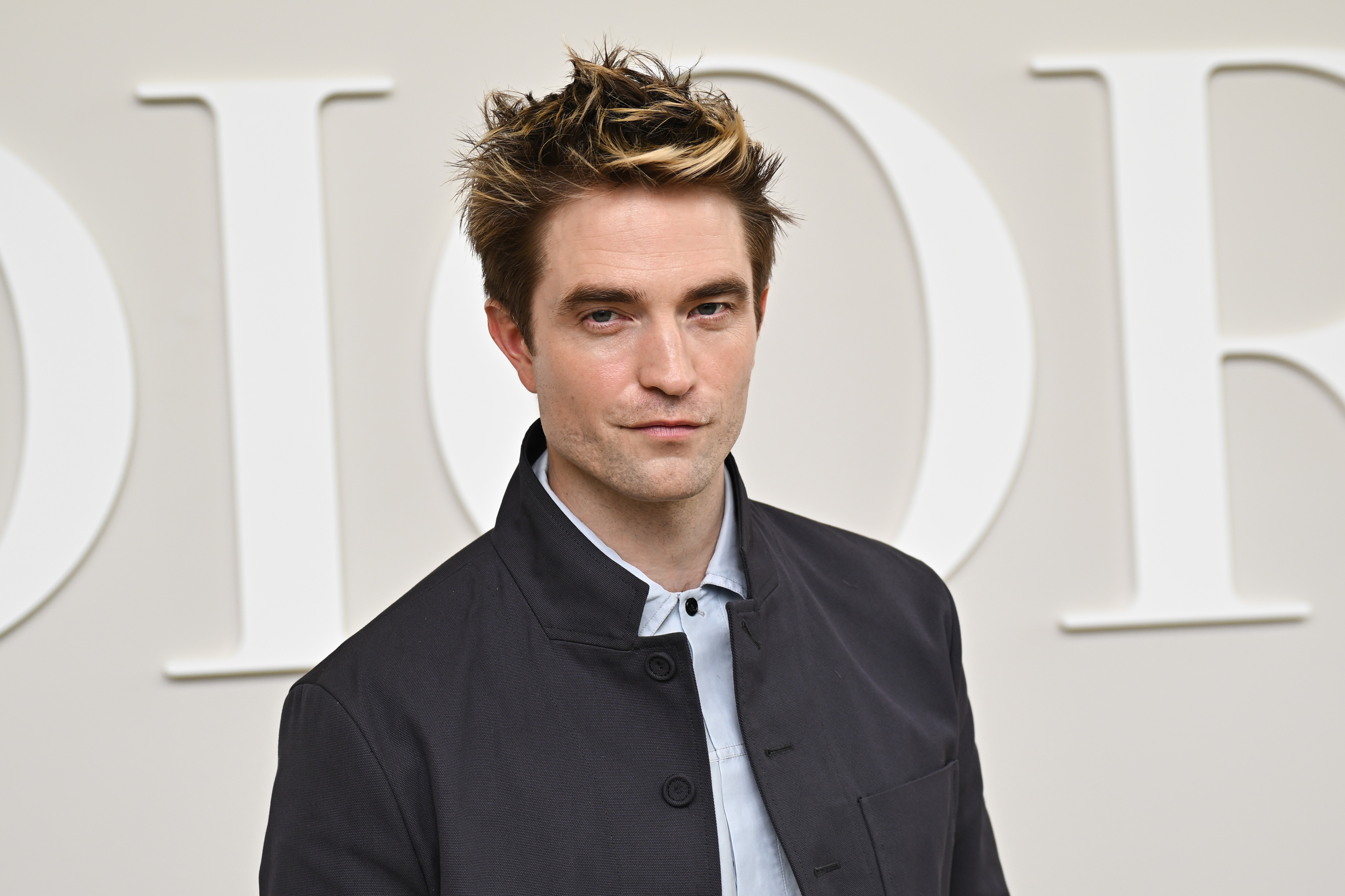 Robert Pattinson poses in a button-up shirt and jacket at an event with large &quot;Dior&quot; signage in the background