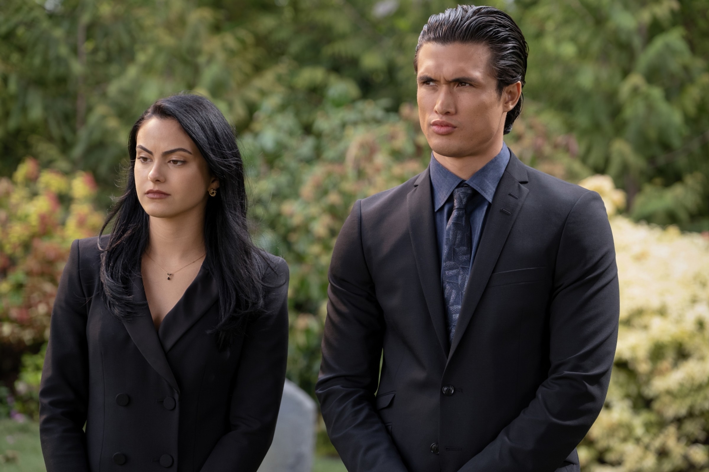 Veronica and Reggie, both dressed in dark, formal suits, stand outside, looking serious