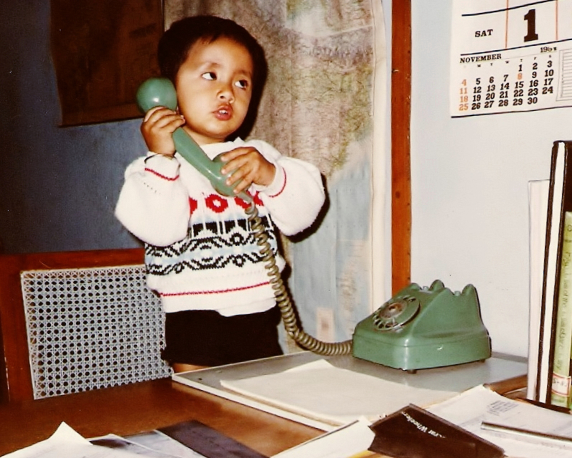 Young child holding a phone receiver to their ear, standing at a desk with papers, a rotary dial phone, and a wall calendar in the background