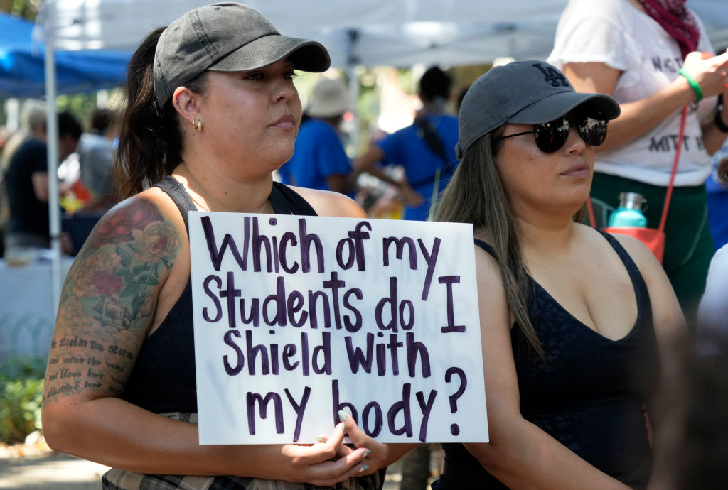 Two women at a protest hold a sign reading, &quot;Which of my students do I shield with my body?&quot; Both wear casual clothing and hats