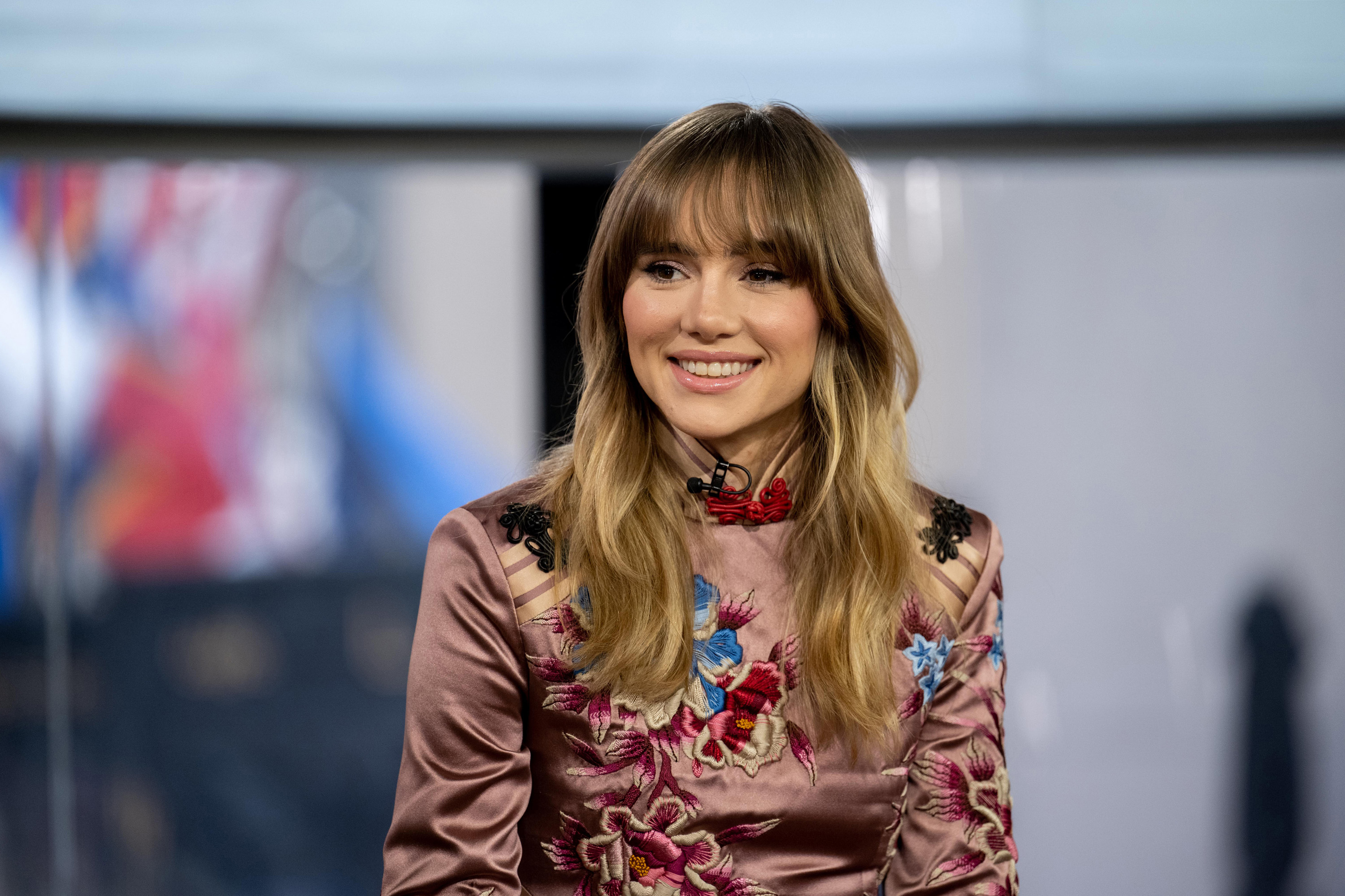 Suki Waterhouse smiles during an interview, wearing a floral patterned satin dress