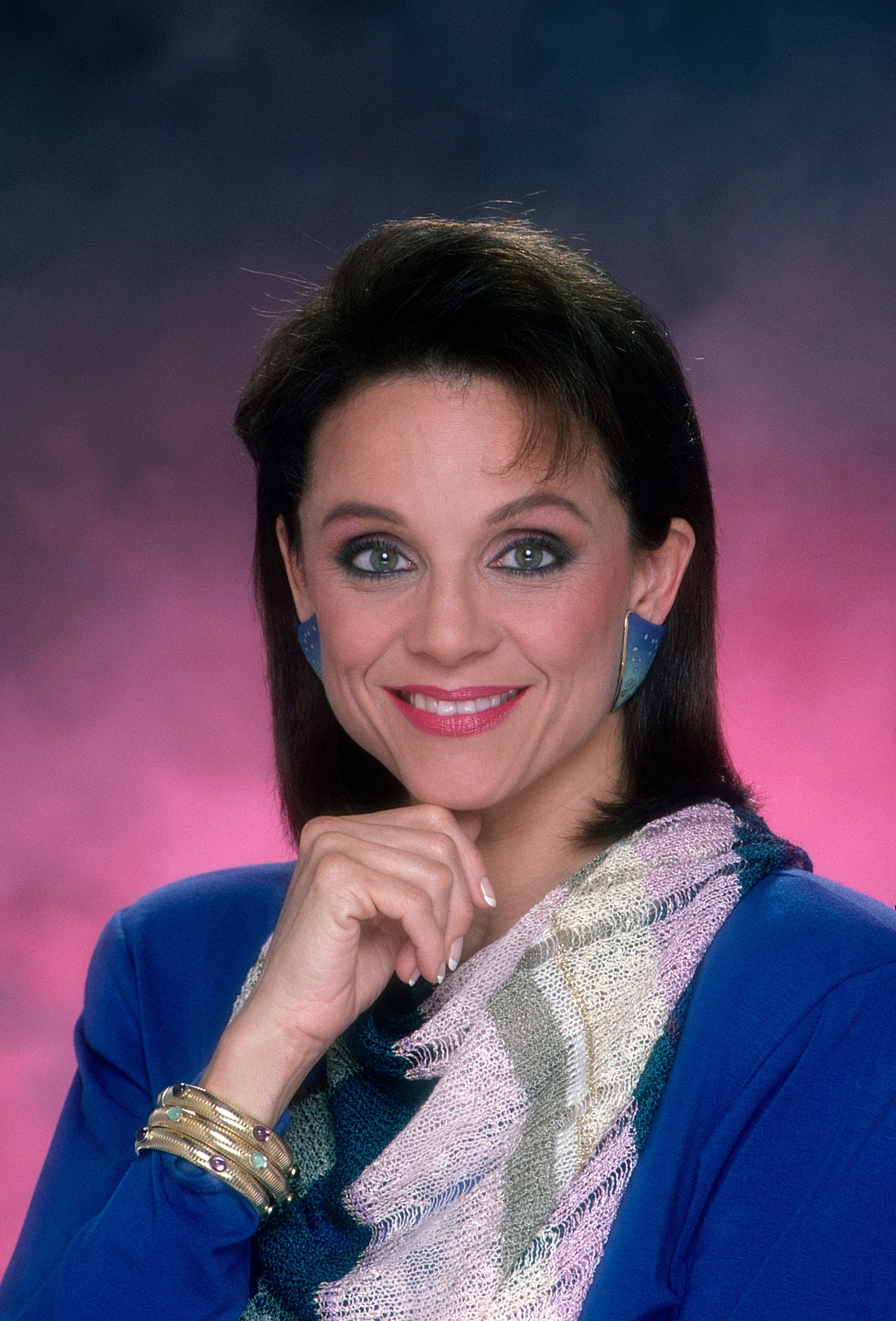 A woman poses with a hand under her chin, smiling, wearing a patterned scarf, bright blue top, and metallic earrings