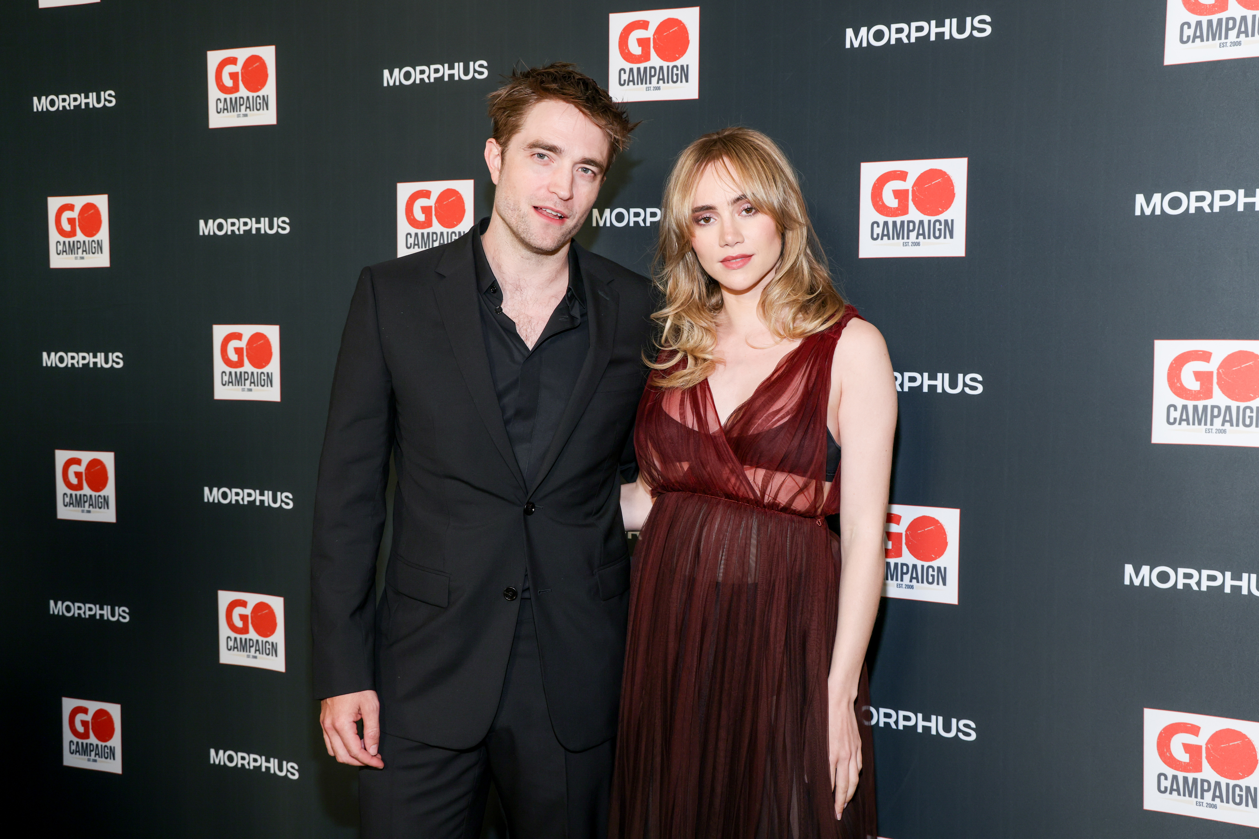 Robert Pattinson and Suki Waterhouse pose together in elegant evening wear at the GO Campaign event backdrop