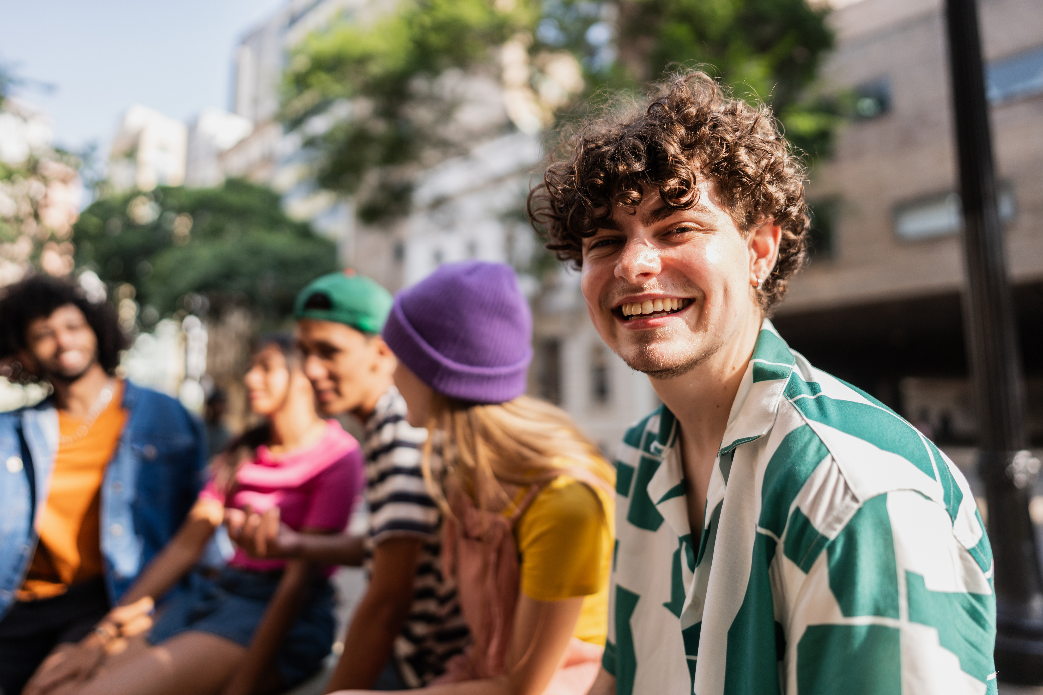 A group of people sit outdoors, smiling and chatting. A curly-haired person in a patterned shirt is in the foreground, with others blurred in the background