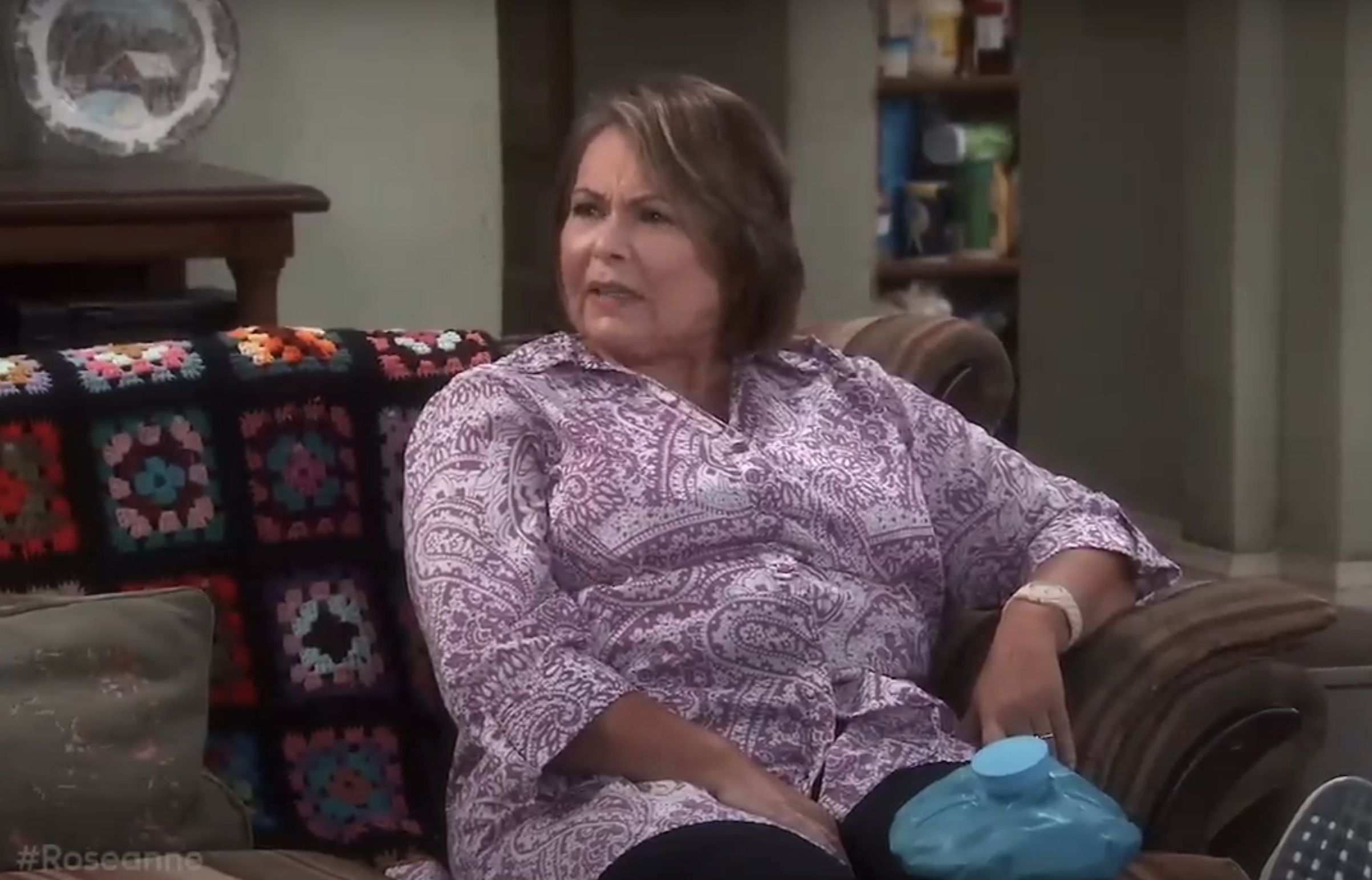 Roseanne Barr sits on a couch, holding an ice pack. The couch has a crocheted blanket on the back. She wears a patterned shirt and appears to be in conversation