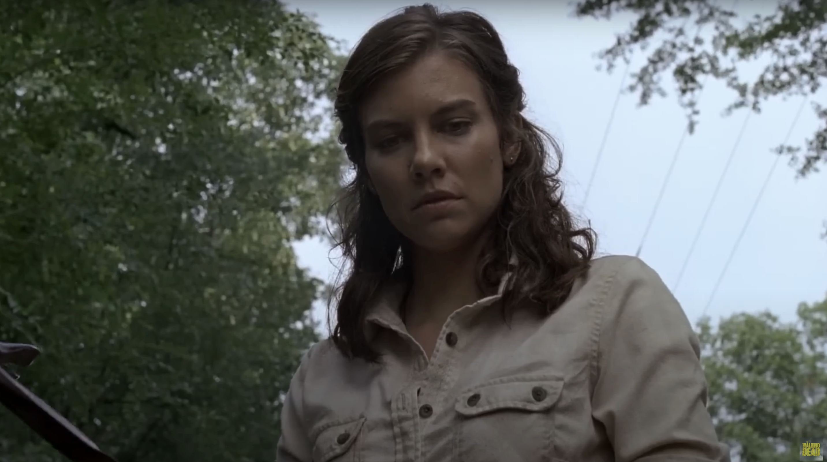 Lauren Cohan in a forest looking down with a serious expression. She wears a buttoned-up shirt