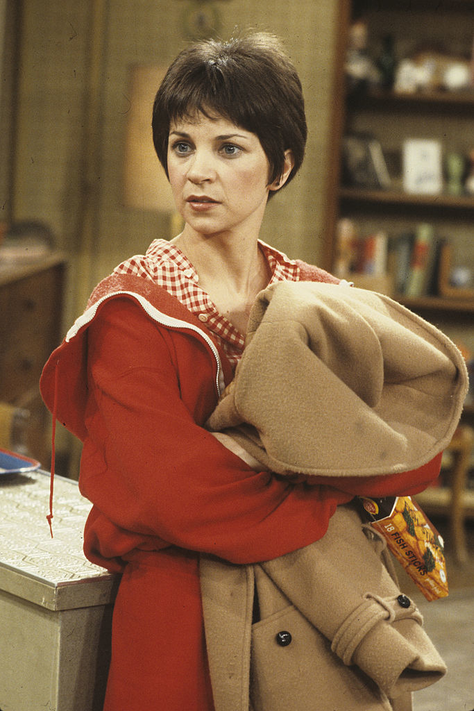 Shirley in jacket with a hood, holding a coat, standing in a room with shelves and books in the background