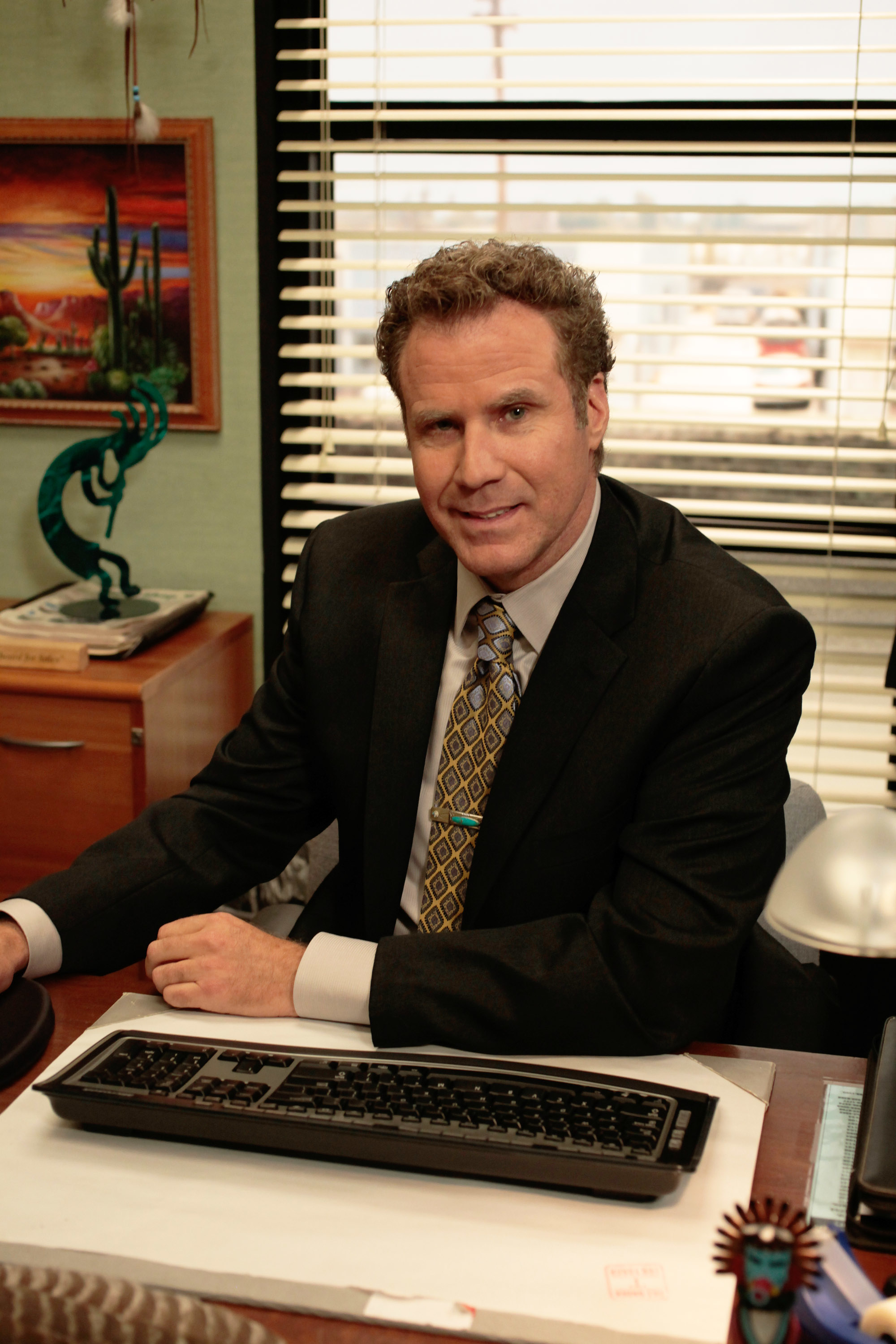Deangelo dressed in a business suit, sitting at a desk in an office setting with a keyboard and various office decor surrounding him