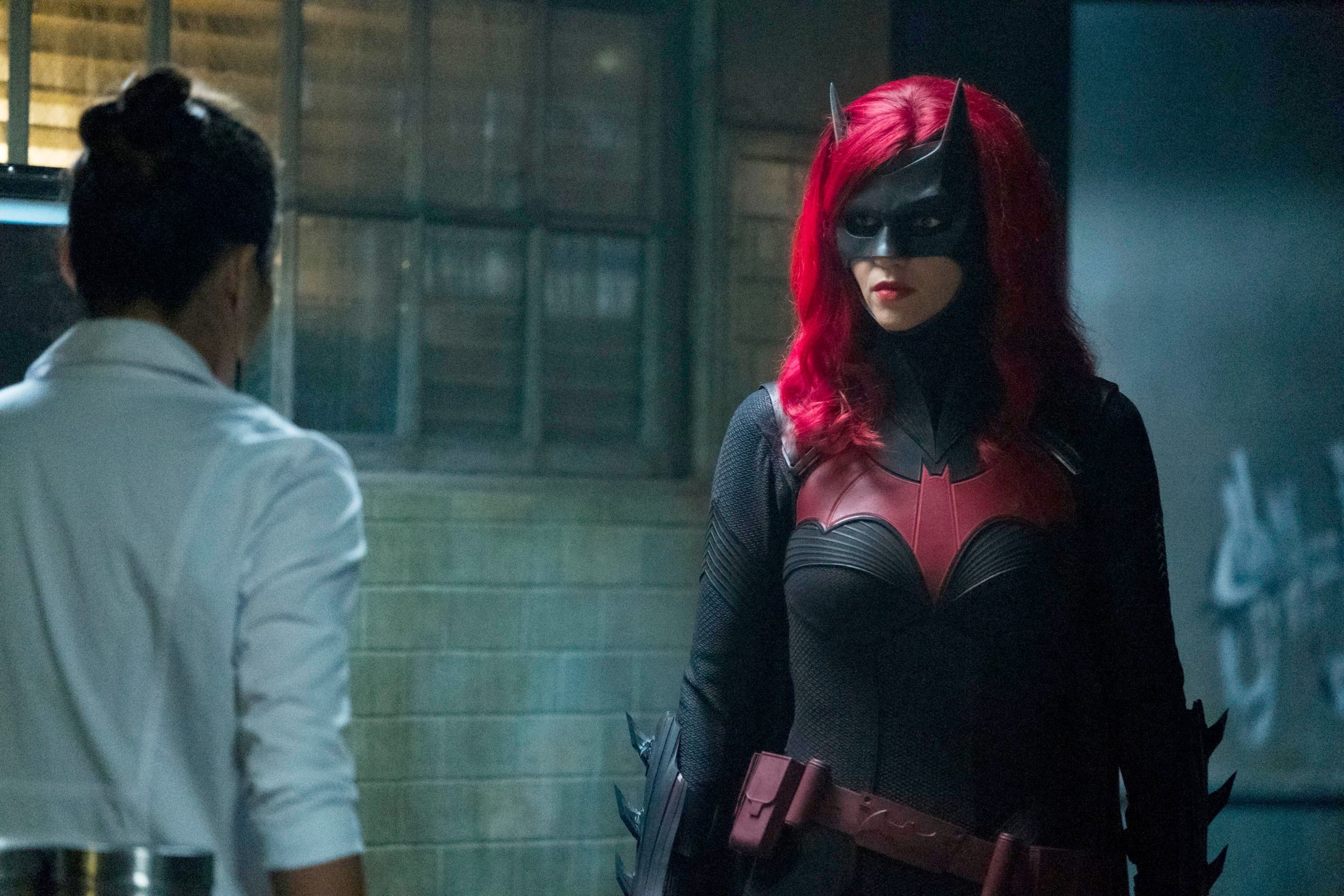 Batwoman, in a dark superhero suit with a bat emblem and bright hair, faces a person with their back turned in a laboratory setting