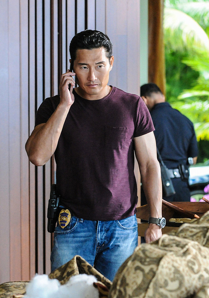 Daniel Dae Kim stands talking on a phone, wearing a casual shirt and jeans with a police badge on his belt. Another officer in uniform is in the background