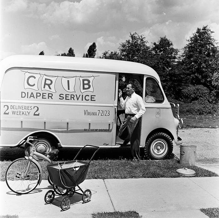 A man stands beside a van labeled &quot;CRIB Diaper Service&quot; holding a phone. A baby carriage and tricycle are in the foreground