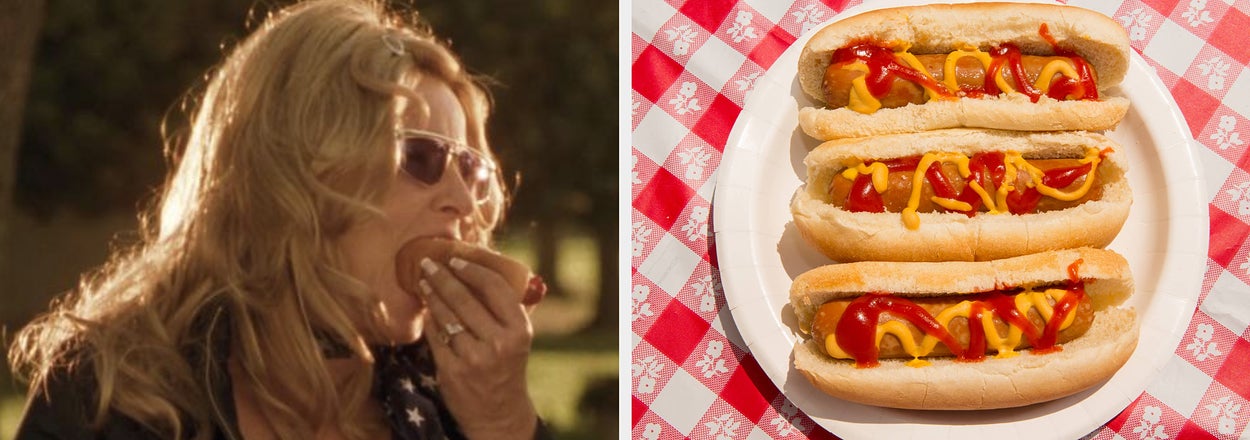 A woman wearing sunglasses is eating a hot dog. Three hot dogs with mustard and ketchup are on a plate with a checkered tablecloth in the background