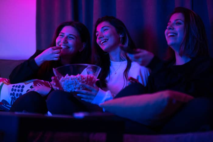 Three women sitting on a couch, smiling and eating popcorn, watching TV