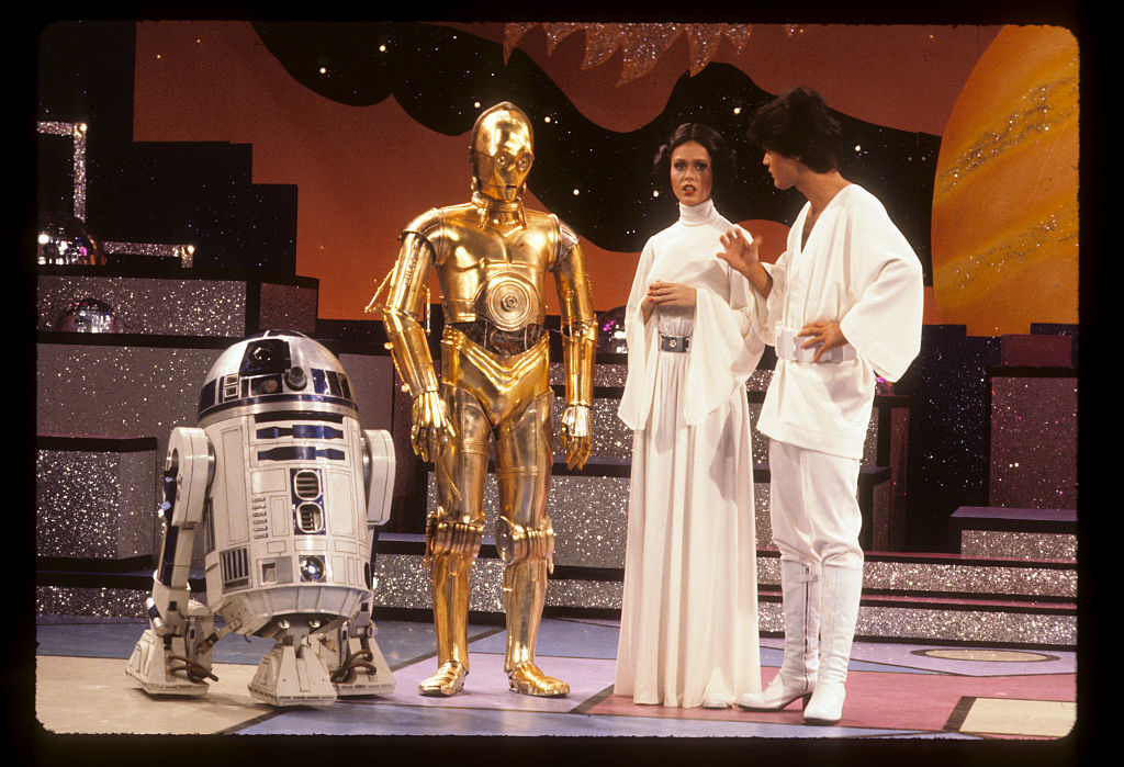 R2-D2, C-3PO, Carrie Fisher, and Mark Hamill are on a stage. Fisher and Hamill are dressed in white, iconic outfits from &quot;Star Wars.&quot;