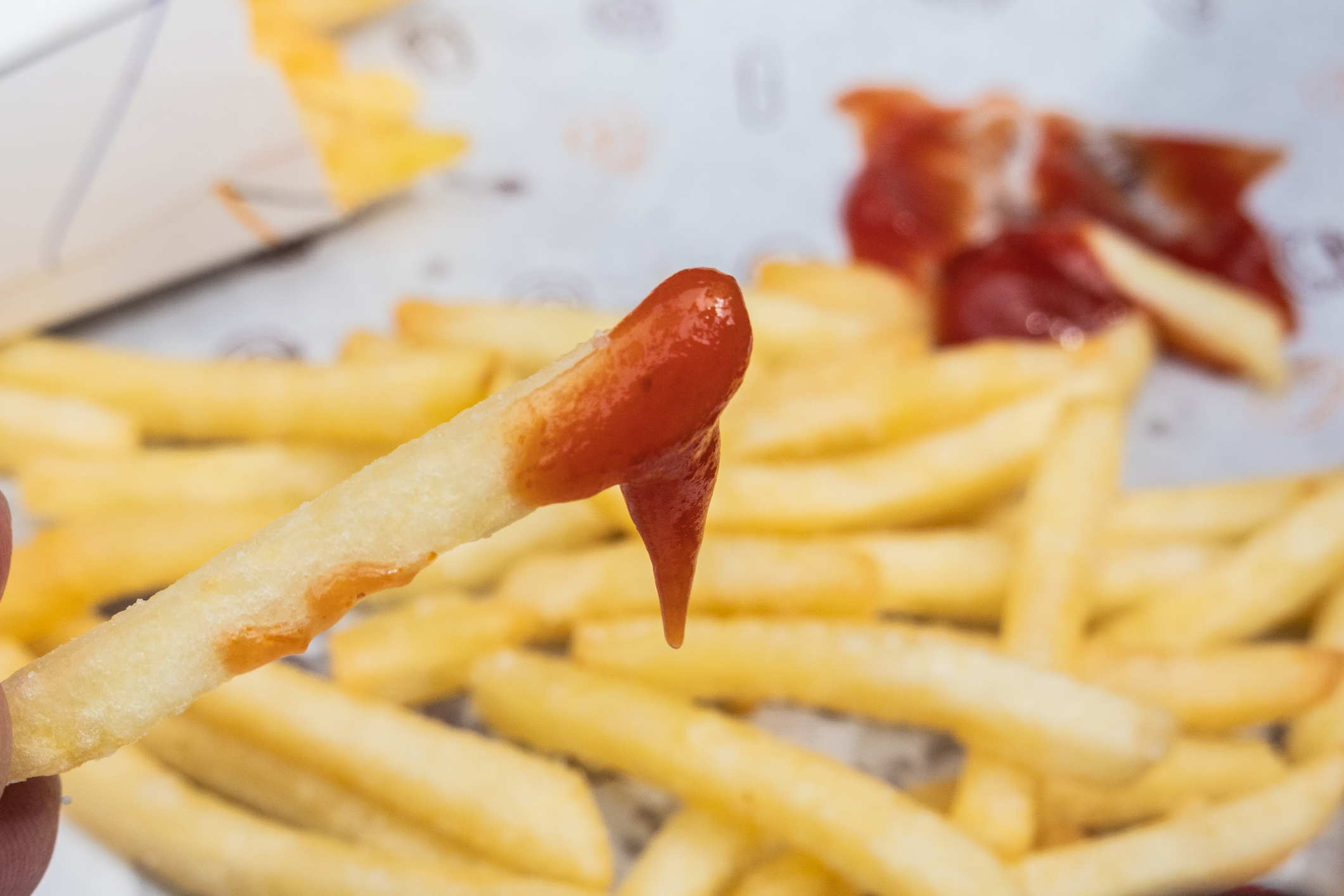 Close-up of a French fry dipped in ketchup, held in front of a pile of fries on a white paper