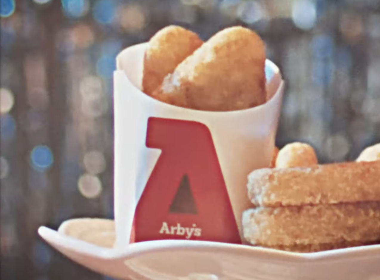 Arby&#x27;s food tray with two mozzarella sticks in a branded container and several onion rings beside it. The setting appears festive with blurred lights