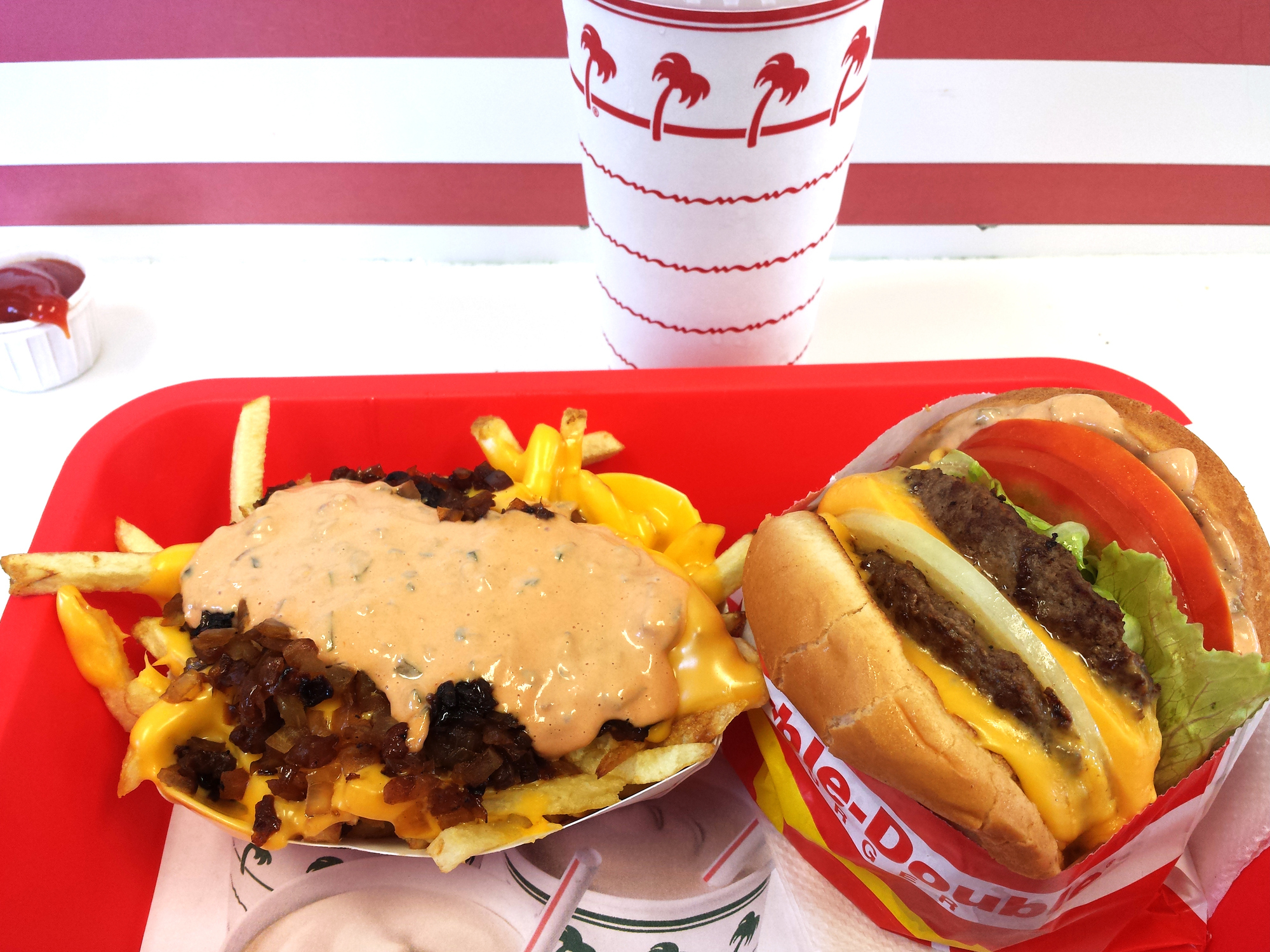 A tray from In-N-Out Burger with Animal Style fries topped with cheese, grilled onions, and sauce, alongside a cheeseburger with lettuce and tomato, and a soda cup