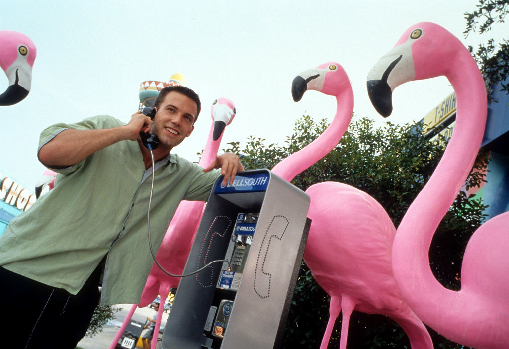 Ben Affleck uses a payphone in an outdoor setting with large pink flamingo statues around him