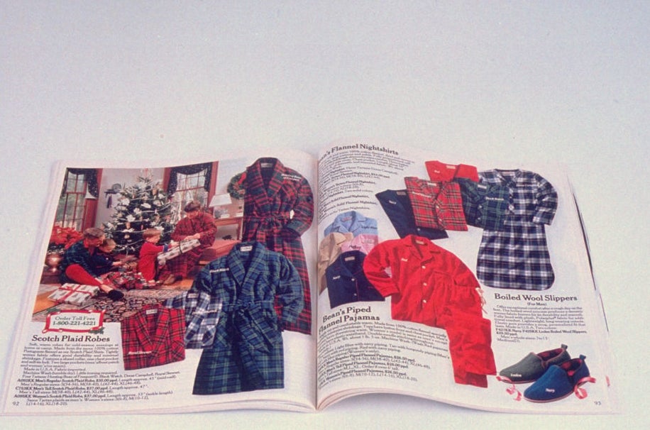 Open catalogue featuring Scotch plaid robes, pajamas, slippers, and various clothing items displayed on pages. The image includes holiday décor in the background