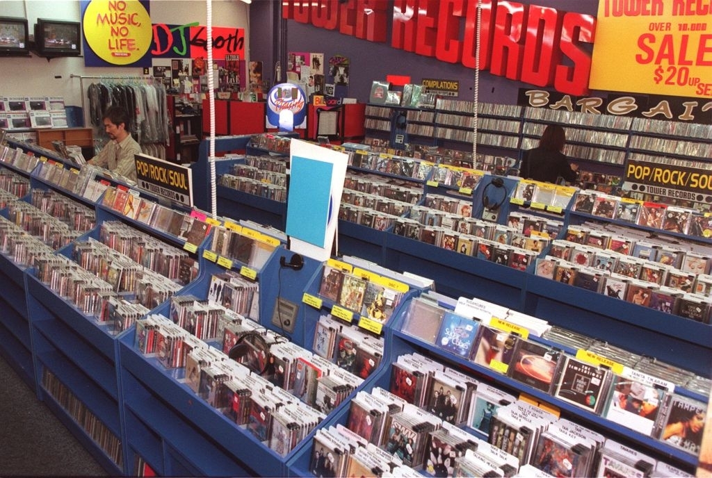 A busy record store with customers browsing CDs and vinyl records. Signs read &quot;Tower Records,&quot; &quot;NO MUSIC NO LIFE,&quot; and &quot;SALE $20up.&quot;