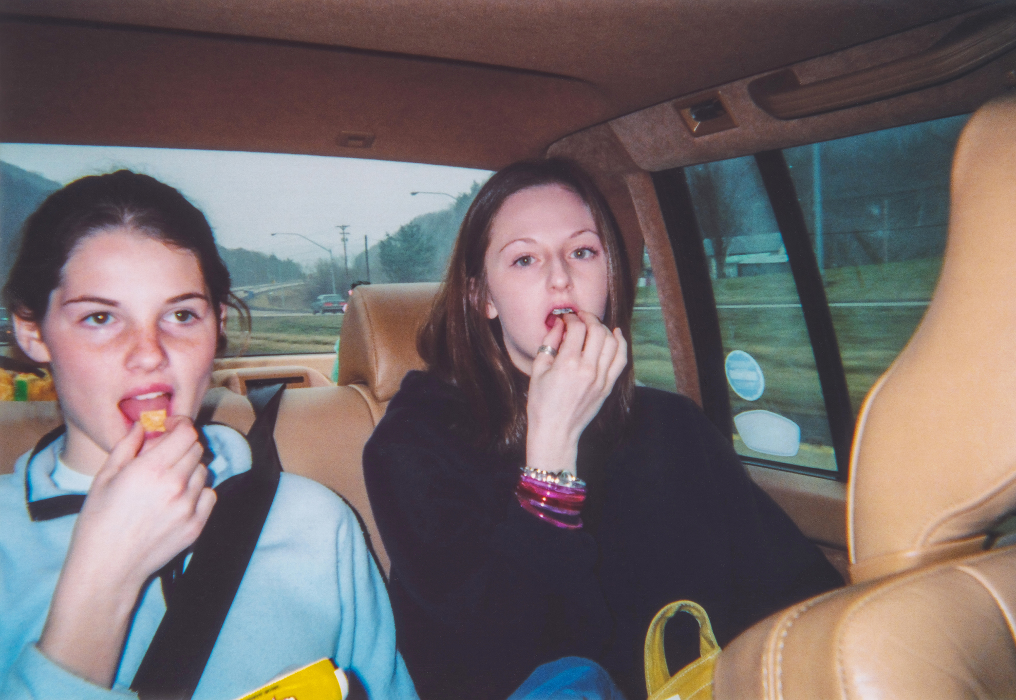 Two teenage girls in casual clothes eating snacks in the backseat of a car
