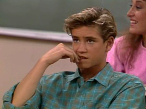 Zack Morris from &quot;Saved by the Bell&quot; sits in a classroom with Kelly Kapowski behind him, smiling