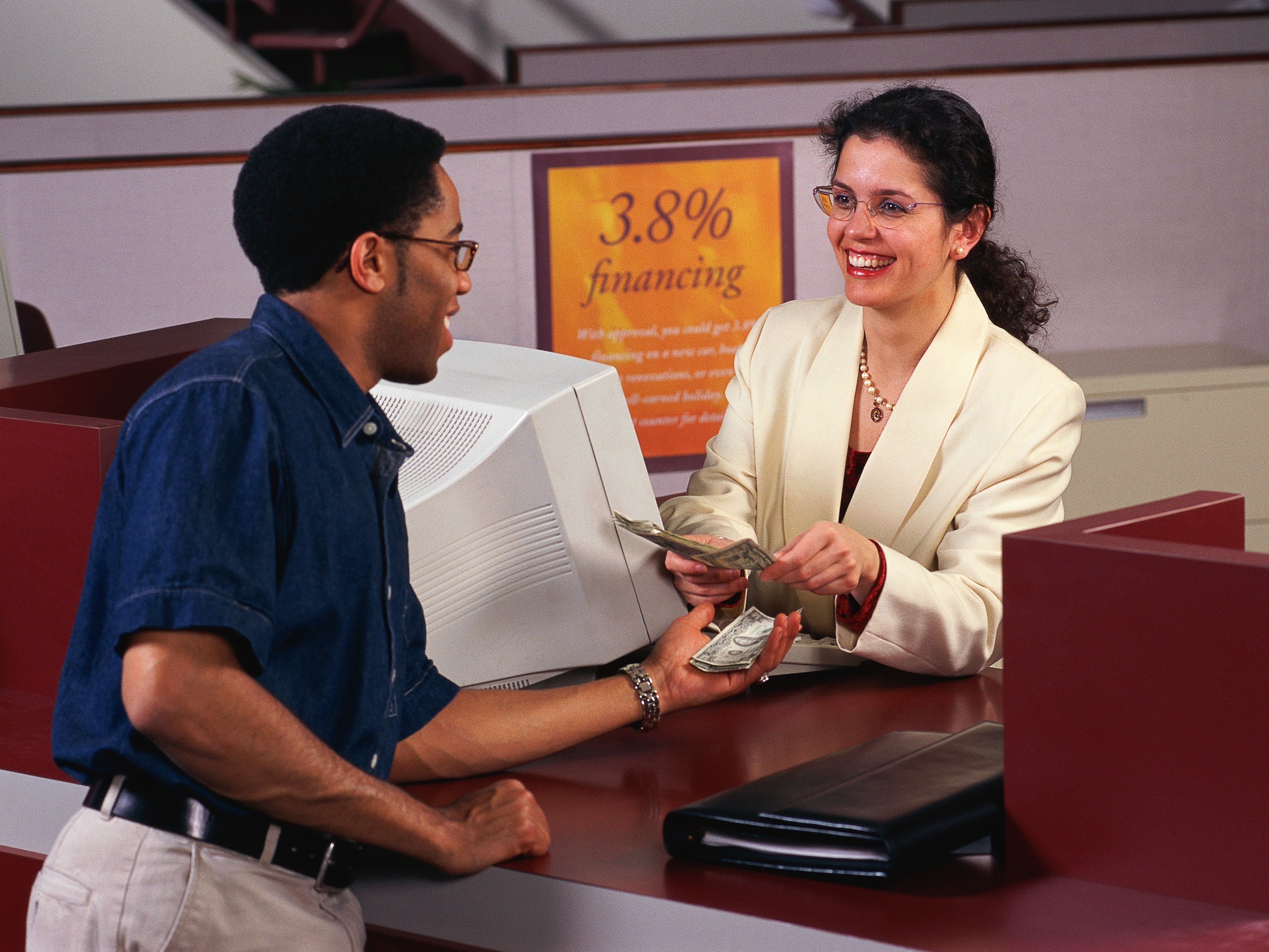 A man in a casual shirt receives cash from a smiling bank teller, who wears a white blazer. A sign in the background advertises 3.8% financing