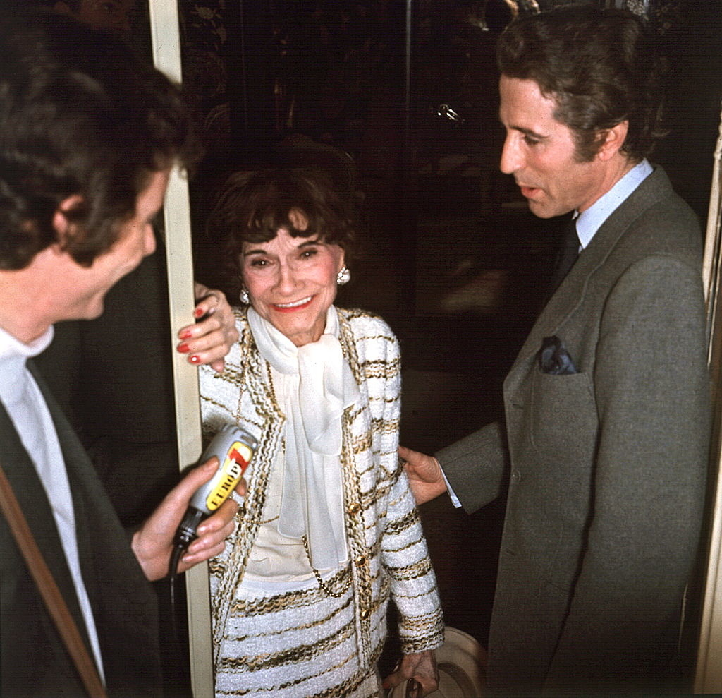 I don&#x27;t know who is in this image. An elderly woman in a striped jacket and white dress is smiling while holding the door, speaking to two men. One man is holding a microphone