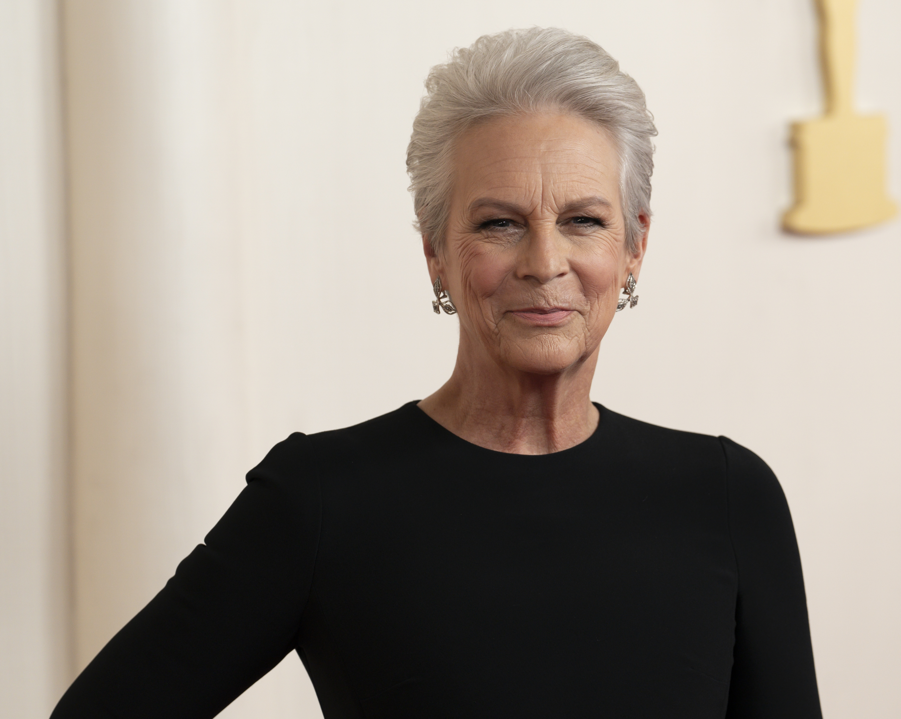 Jamie Lee Curtis stands in front of an Oscar statue, wearing a sleek, simple black dress with her hand on her hip, and sporting short hair