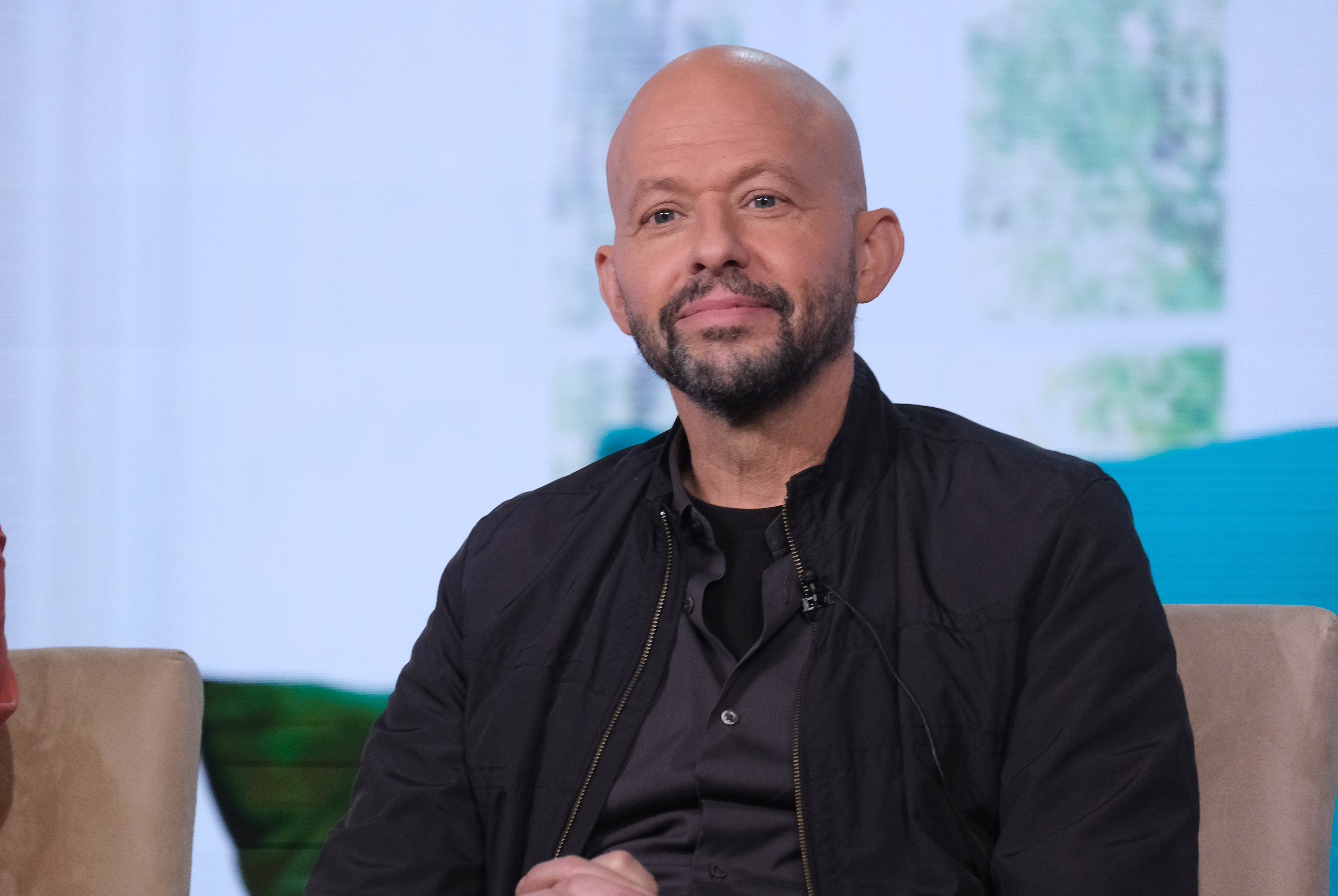 Jon Cryer is seated, wearing a black jacket over a dark shirt, looking ahead with a slight smile