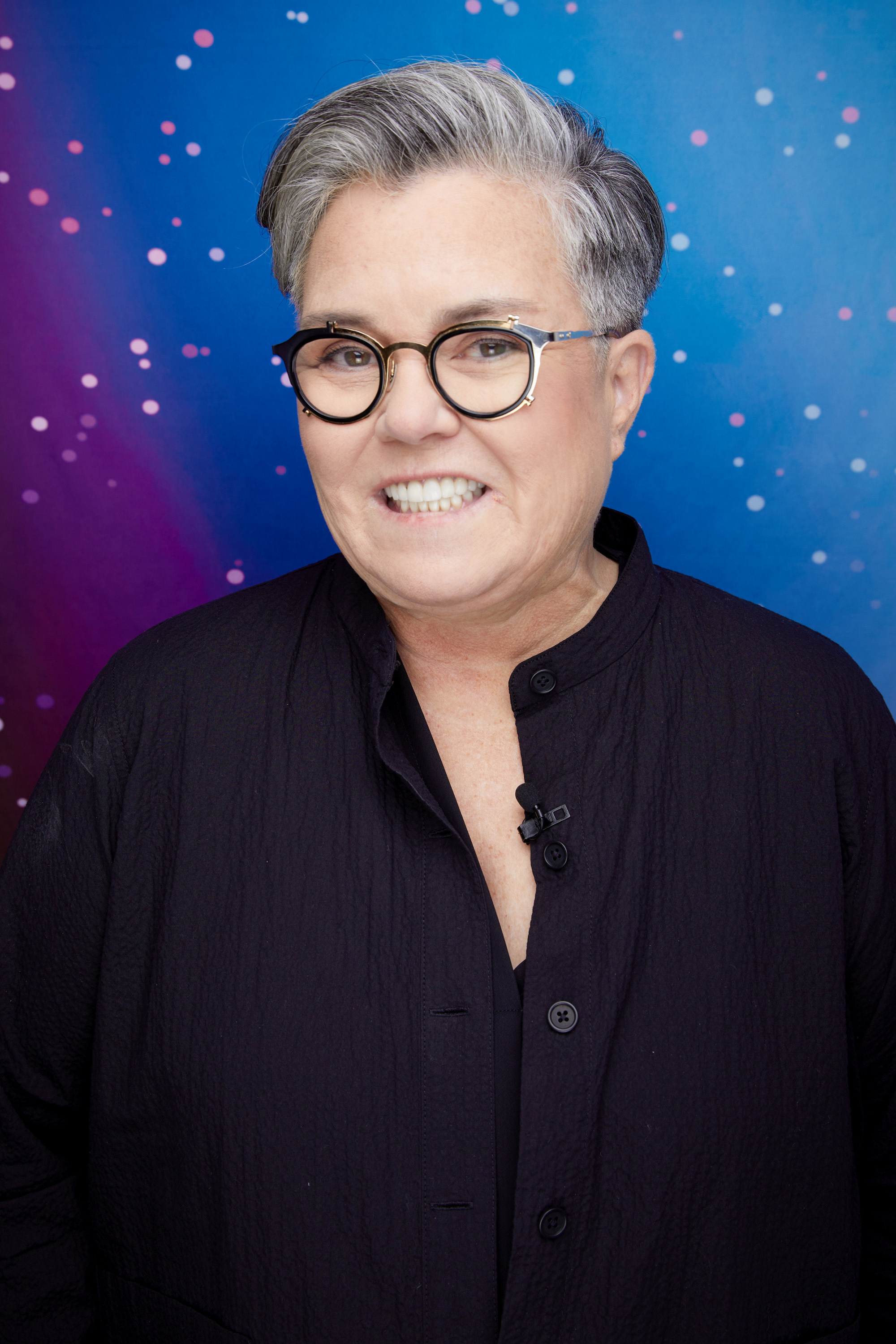 Rosie O&#x27;Donnell smiles while wearing a dark-buttoned top and black glasses, against a starry night background