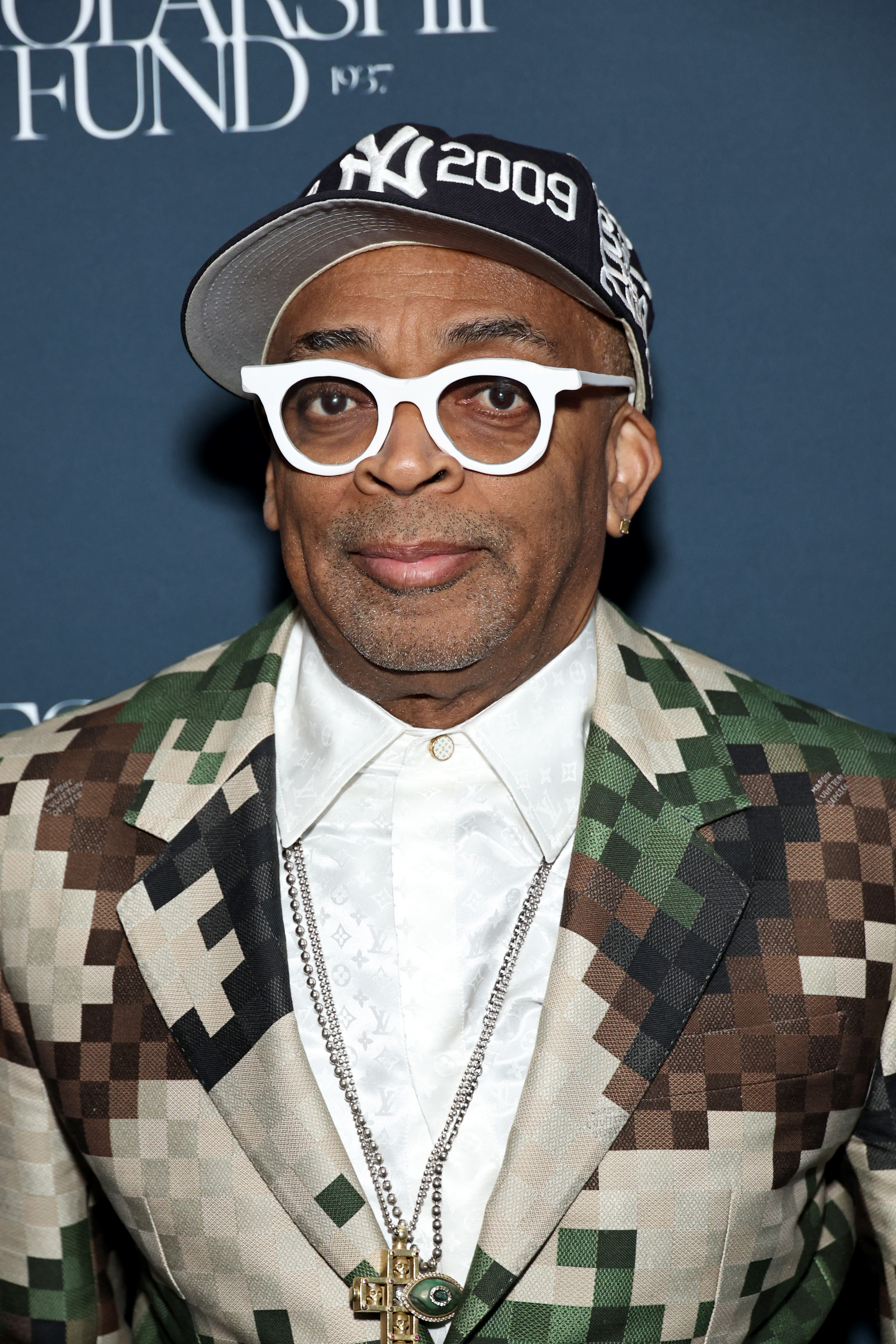 Spike Lee at an event wearing a patterned suit jacket, white shirt, white frame glasses, multiple chains, and a navy baseball cap