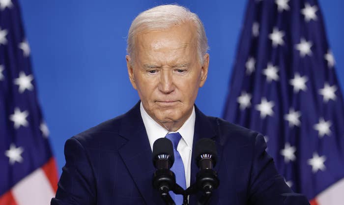 Joe Biden, wearing a suit and tie, is speaking at a podium with microphones in front of U.S. flags