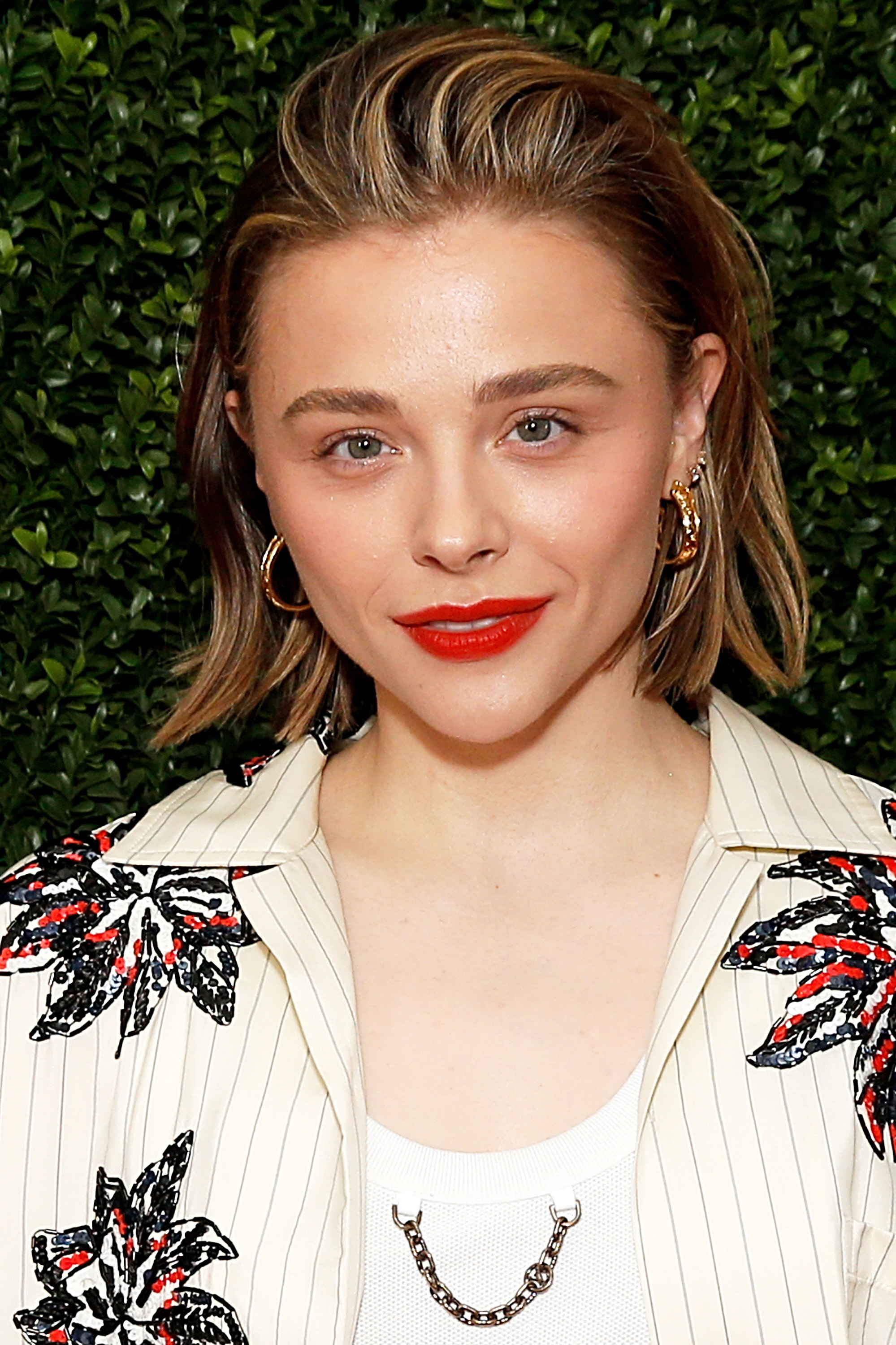 Chloë Grace Moretz smiles in a floral-patterned blazer and a white top with a chain necklace, standing in front of green foliage