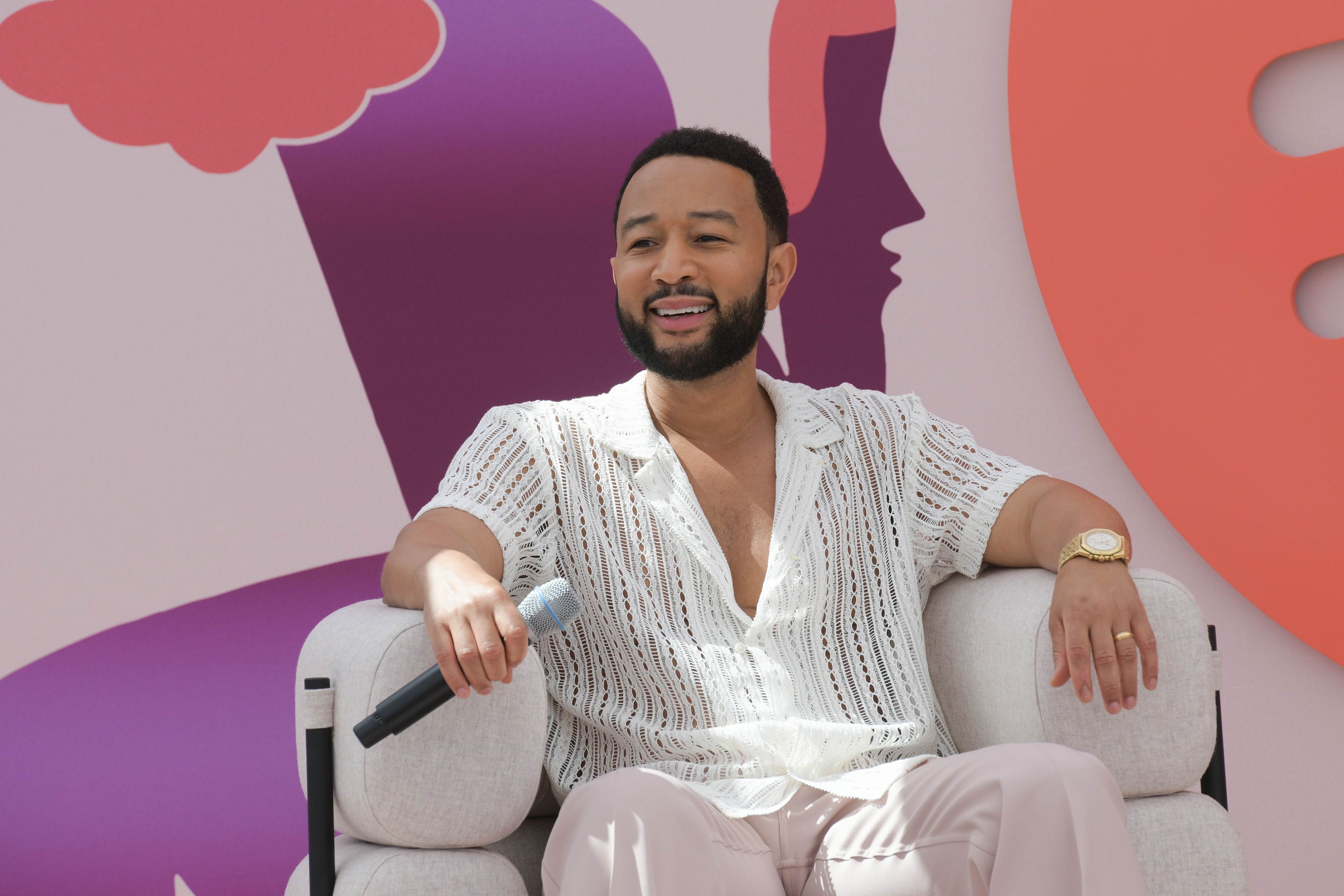 John Legend sits casually on a chair, holding a microphone, wearing a stylish crochet shirt at a public event on stage