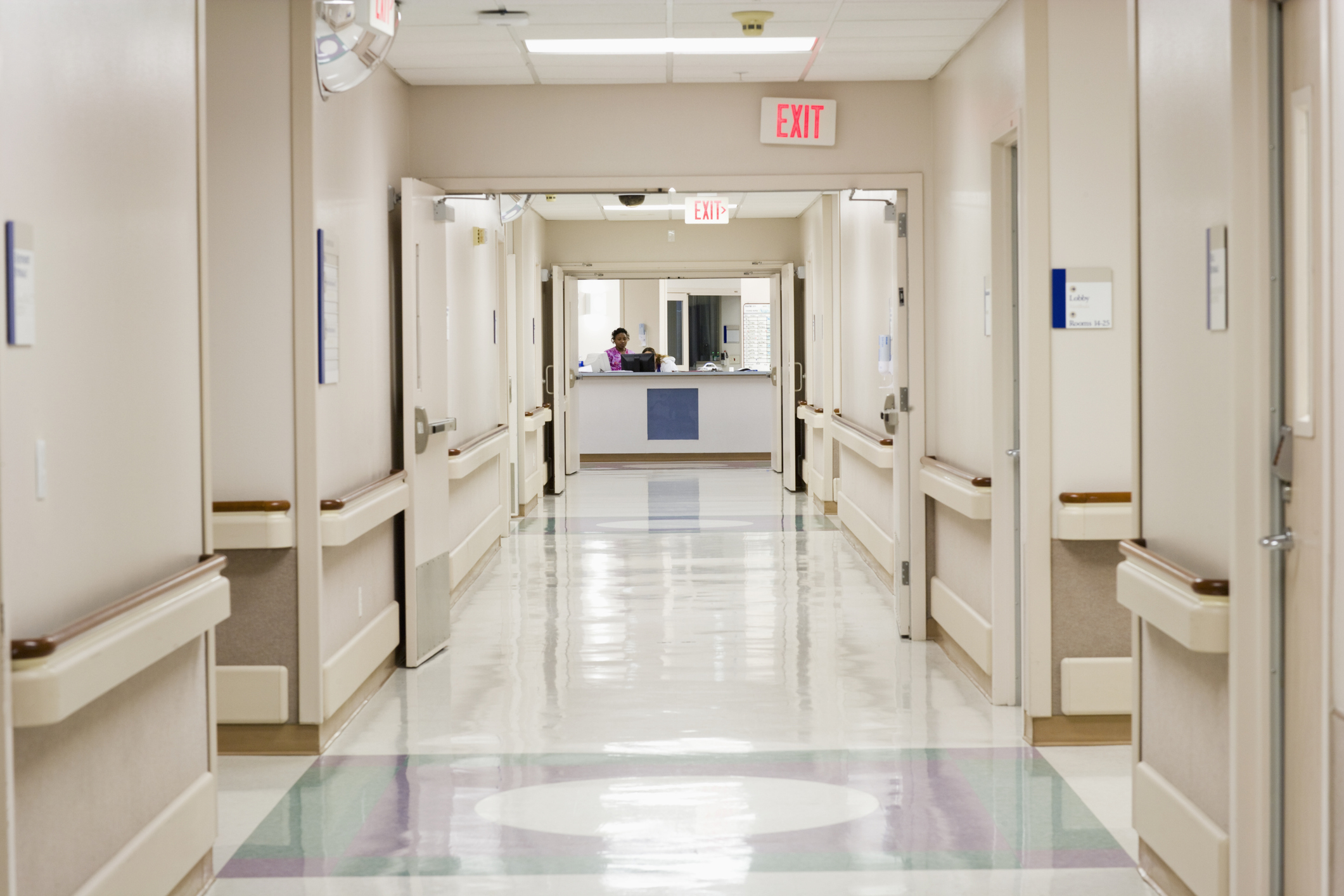 A quiet hospital hallway with shiny floors, handrails, and an exit sign in the background. Two healthcare workers are seen at the nurse&#x27;s station at the end of the hall