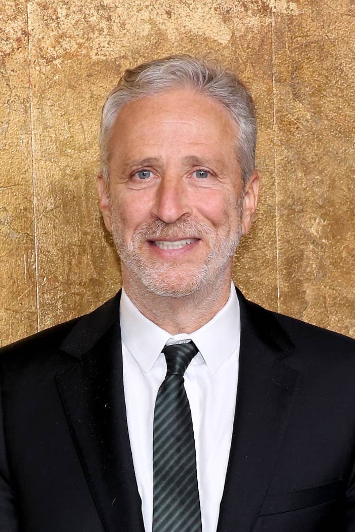 Jon Stewart smiling at an event, wearing a black suit, white shirt, and dark tie against a textured background