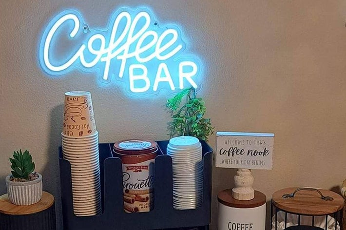 Coffee station setup with a neon "Coffee Bar" sign, paper cups, a tin of cocoa, plants, and a "Welcome to the coffee nook" sign