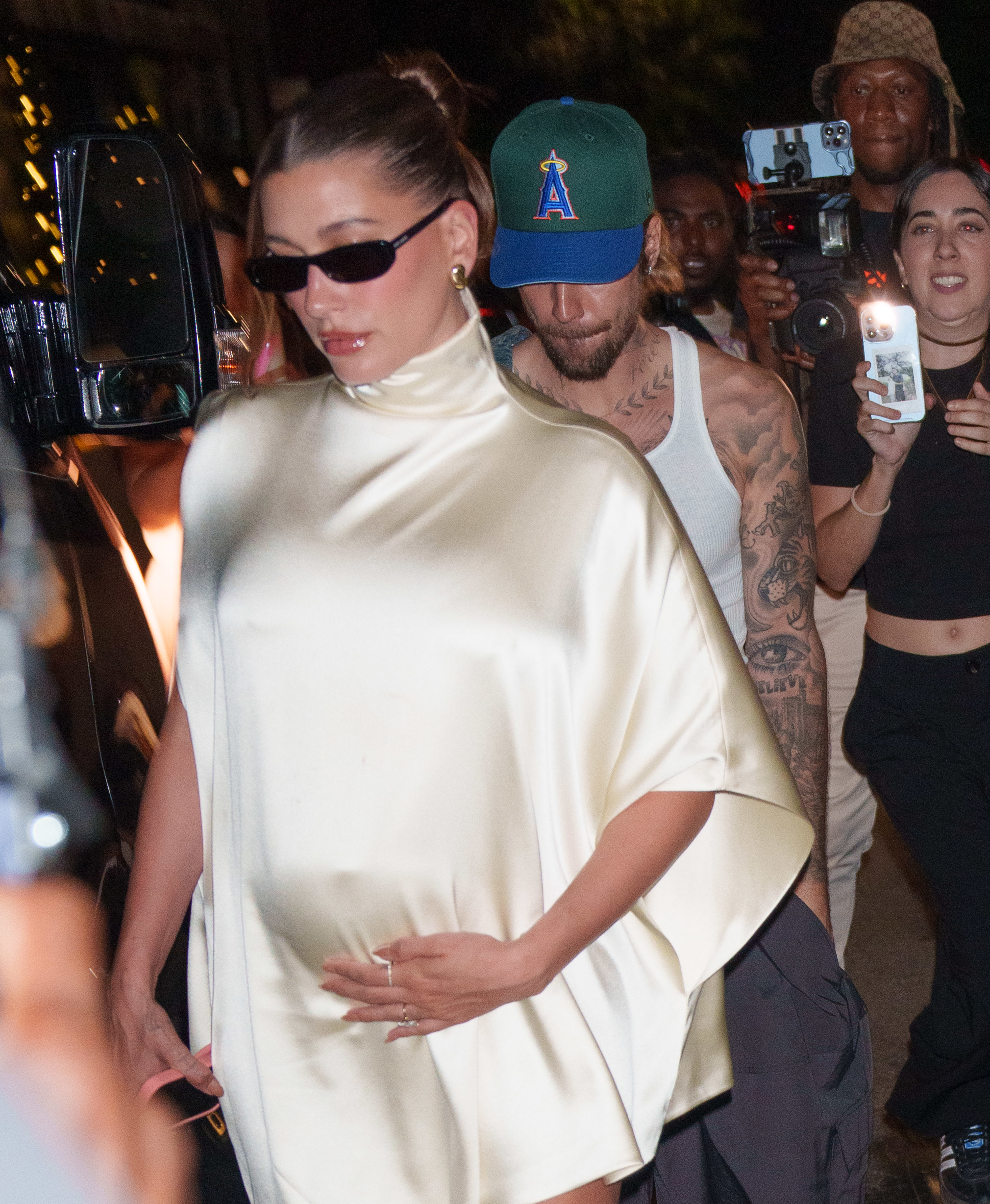 Hailey Bieber and Justin Bieber leaving an event; Hailey wears a stylish, cape-like top, and Justin is behind her in a tank top and cap