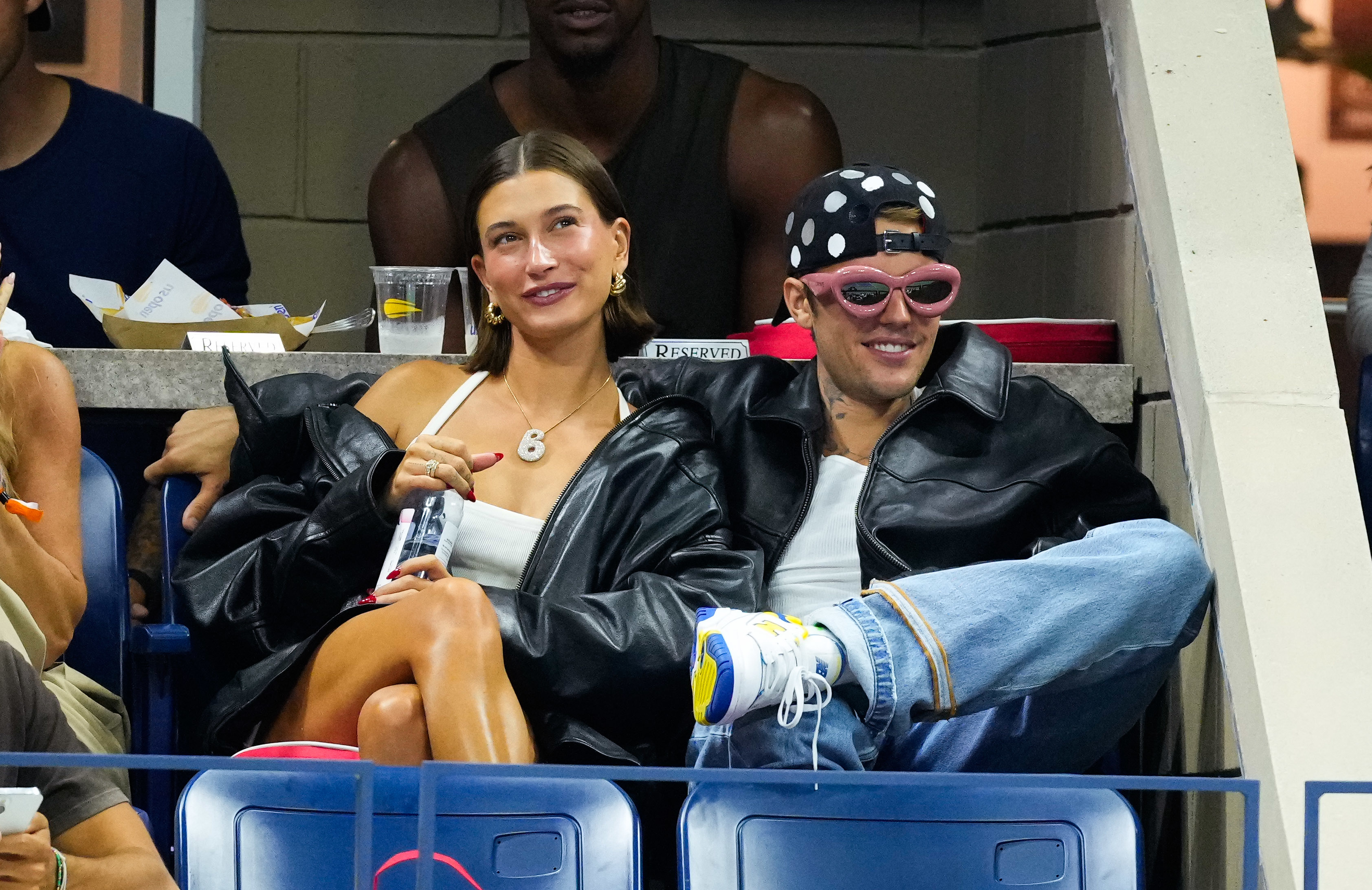 Hailey Bieber and Justin Bieber sit together at an event, both wearing black jackets. Justin sports a polka dot hat and sunglasses, while Hailey smiles holding a drink