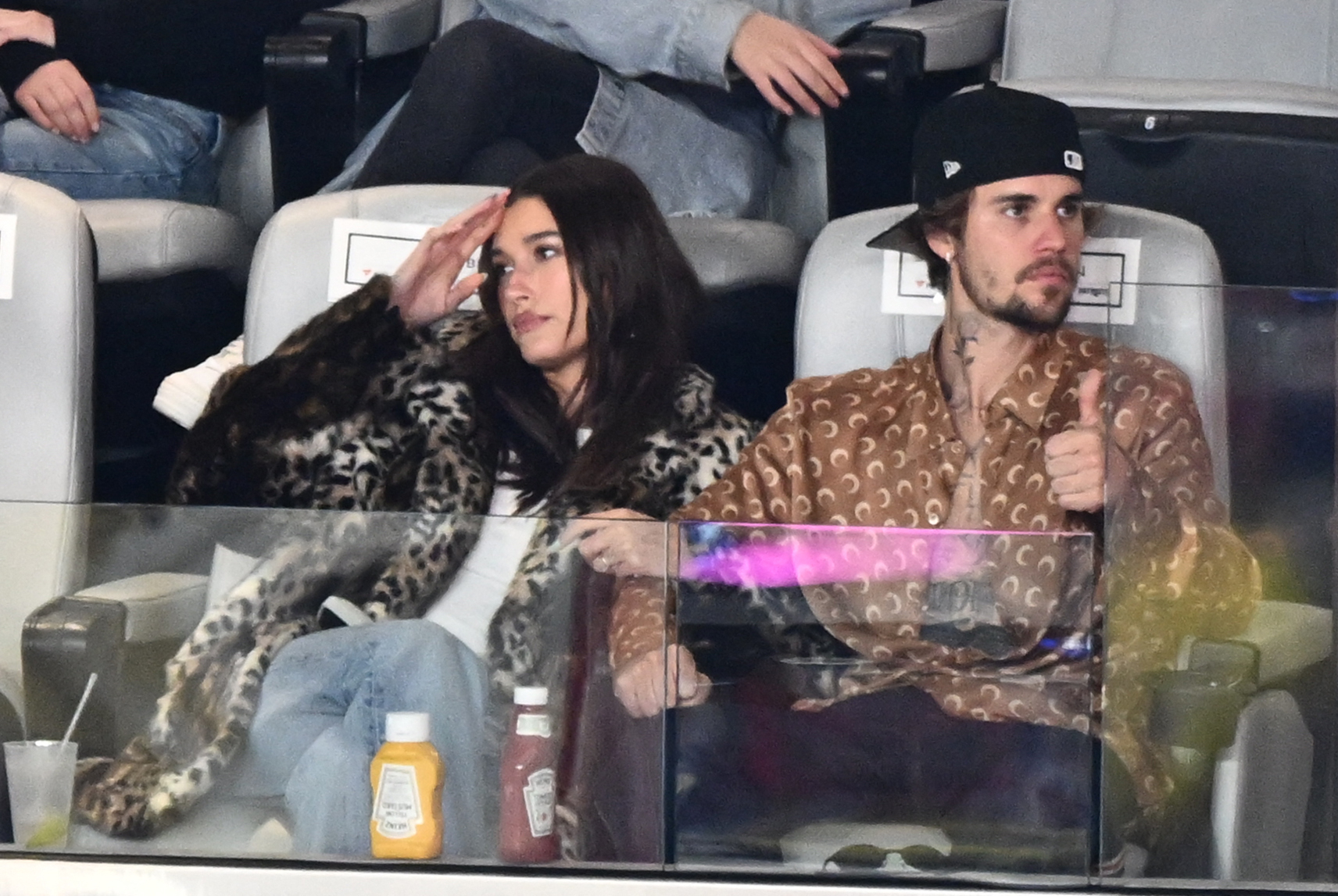 Hailey Bieber in a patterned coat and jeans, and Justin Bieber in a patterned shirt and cap as they sit in a stadium